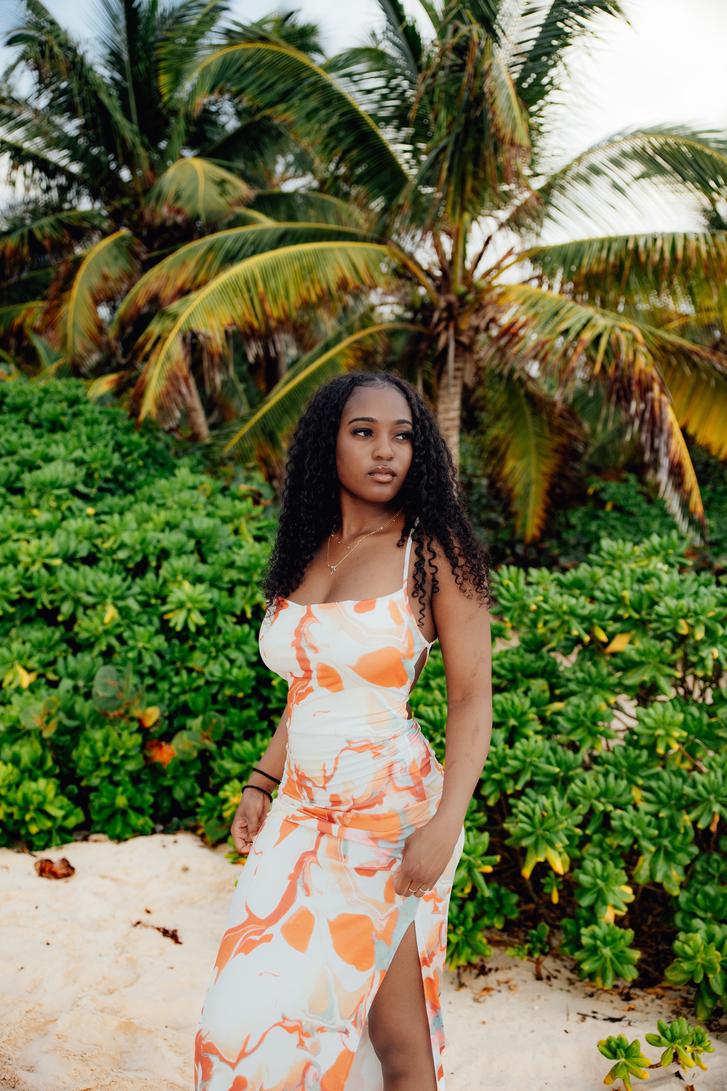 A young woman with dark, curly hair wearing a sleeveless, white and orange dress with a thigh-high slit, standing on a sandy beach in front of lush green bushes and a palm tree.