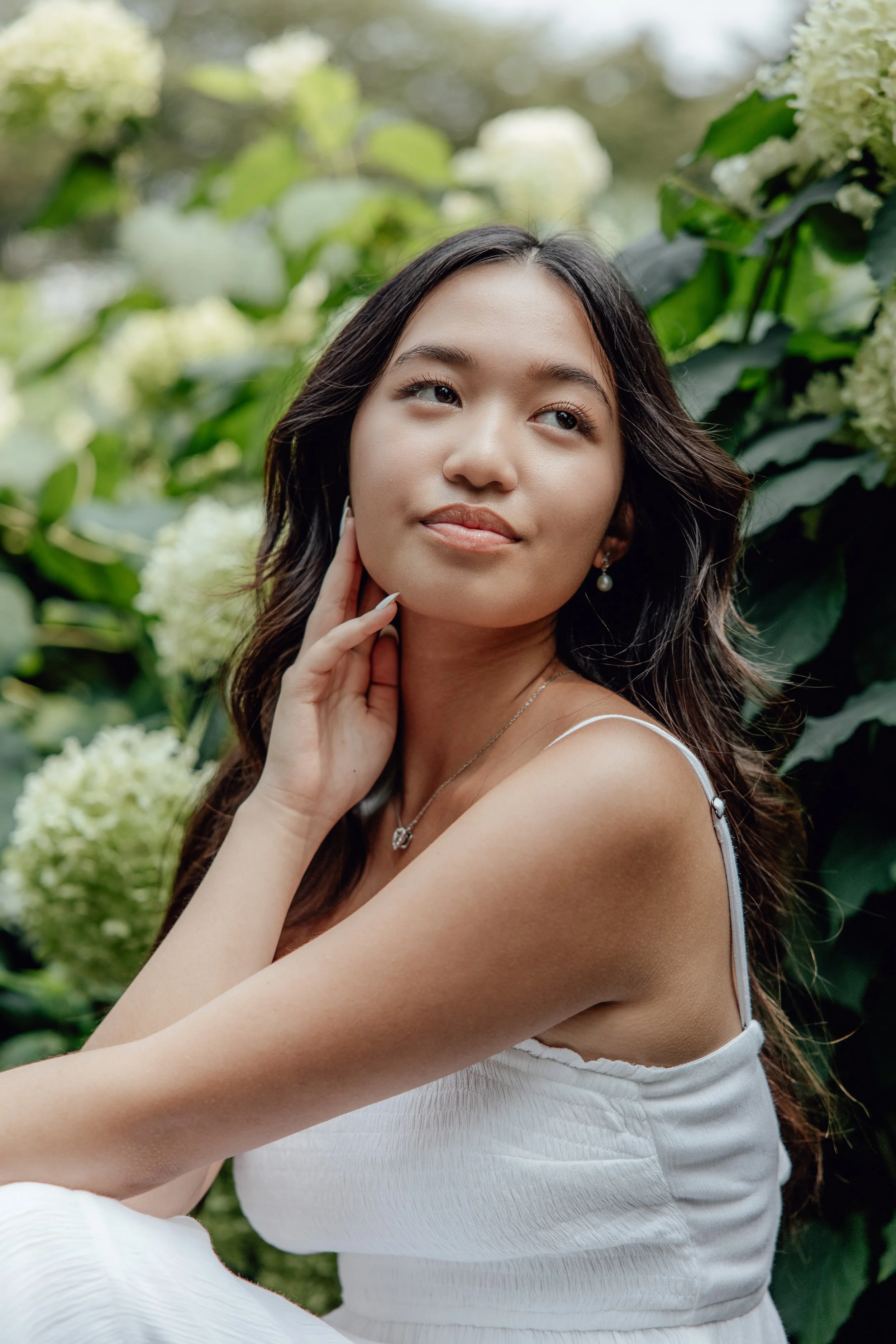 A young Asian woman with long dark hair, wearing a white dress and pearl earrings, is posing outdoors among green foliage and white hydrangea flowers.