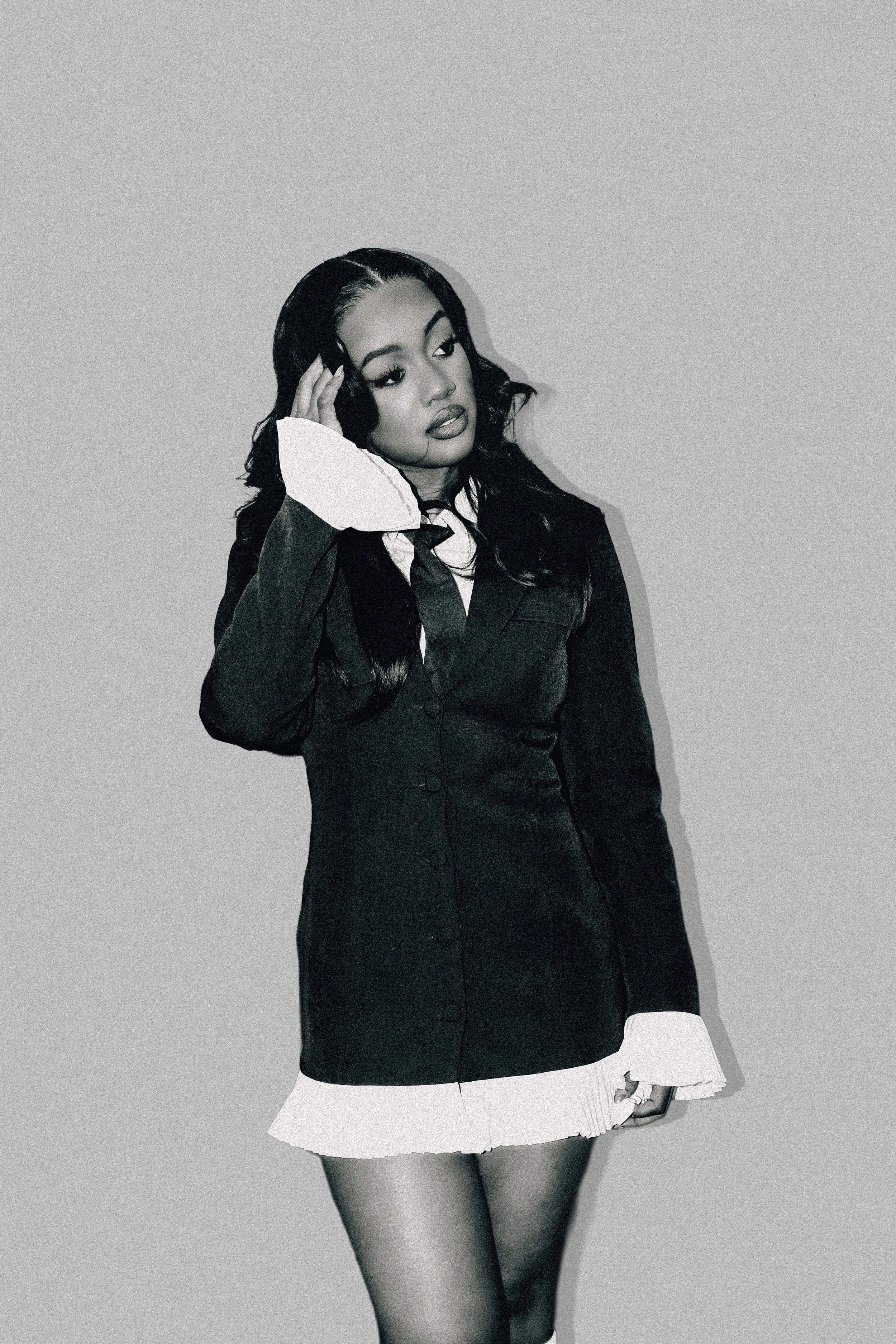 A woman in a black blazer dress with white cuffs and a pleated white hem, wearing black tights and black shoes, posing against a plain background.
