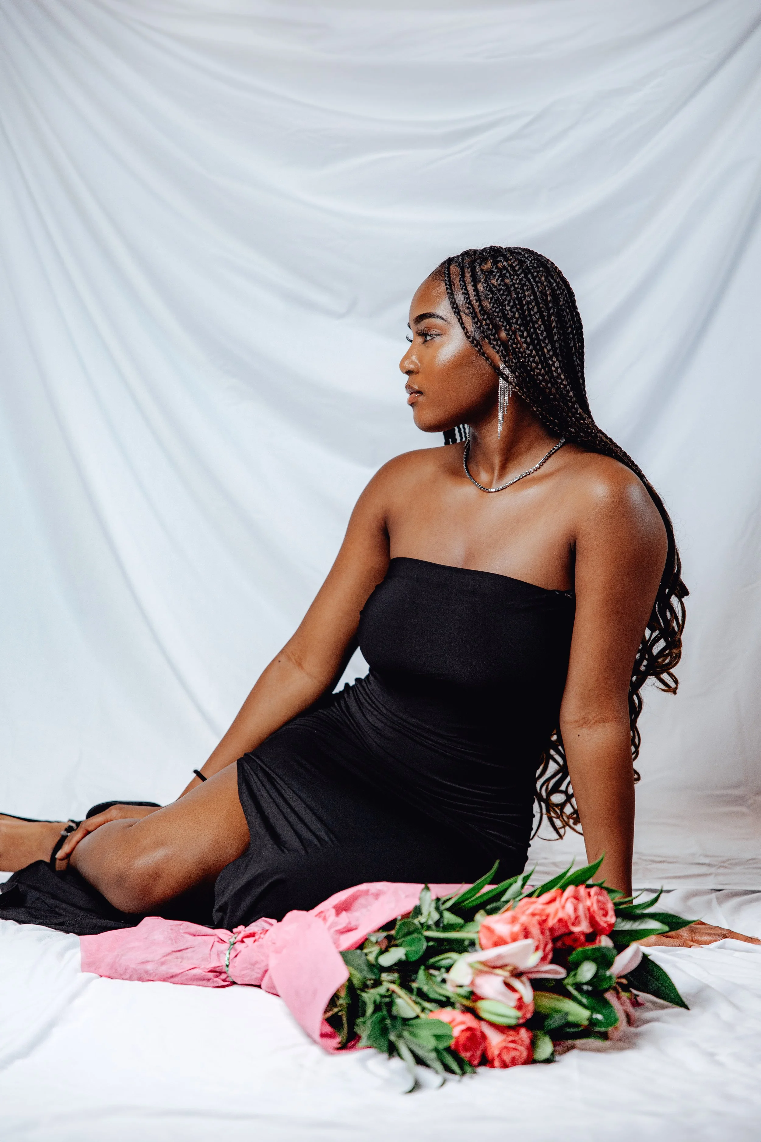 A woman sitting on the floor wearing a black strapless dress, with a bouquet of pink and white flowers wrapped in pink paper in front of her, against a white draped fabric background.
