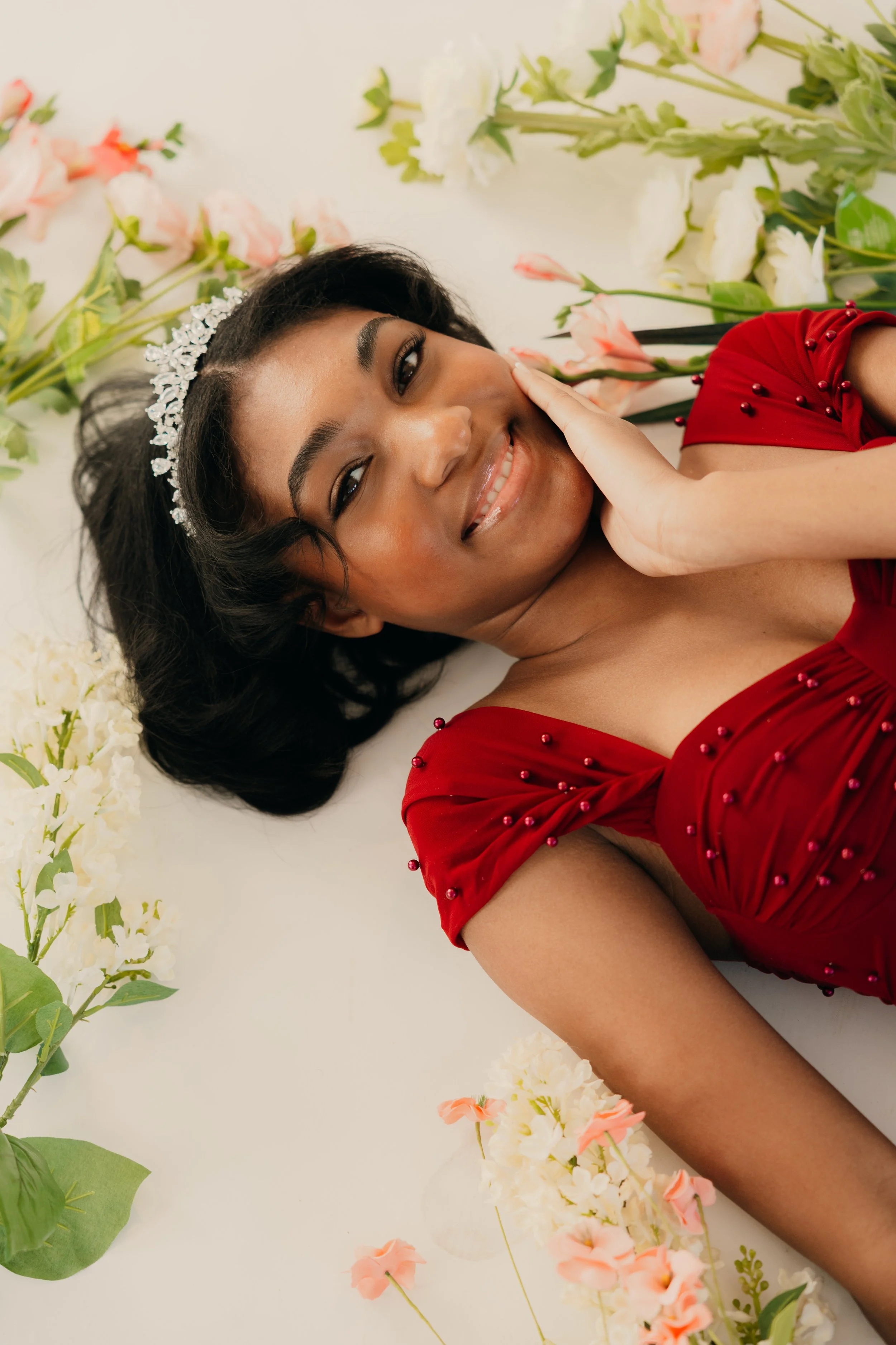 A young woman with dark hair wearing a sparkly tiara and a red dress with beads, smiling and lying on a white surface surrounded by white and pink flowers.