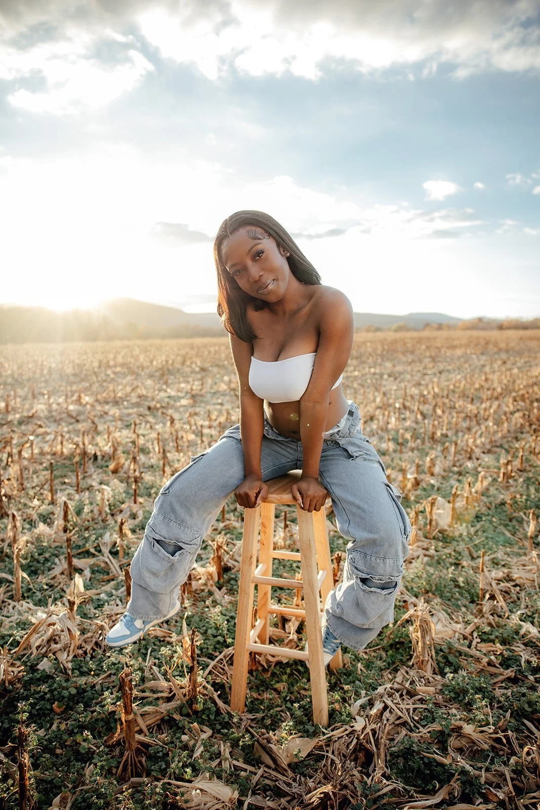 A woman with dark hair and medium skin tone sitting barefoot on a wooden stool in a dried cornfield at sunset, wearing a white crop top and ripped jeans.