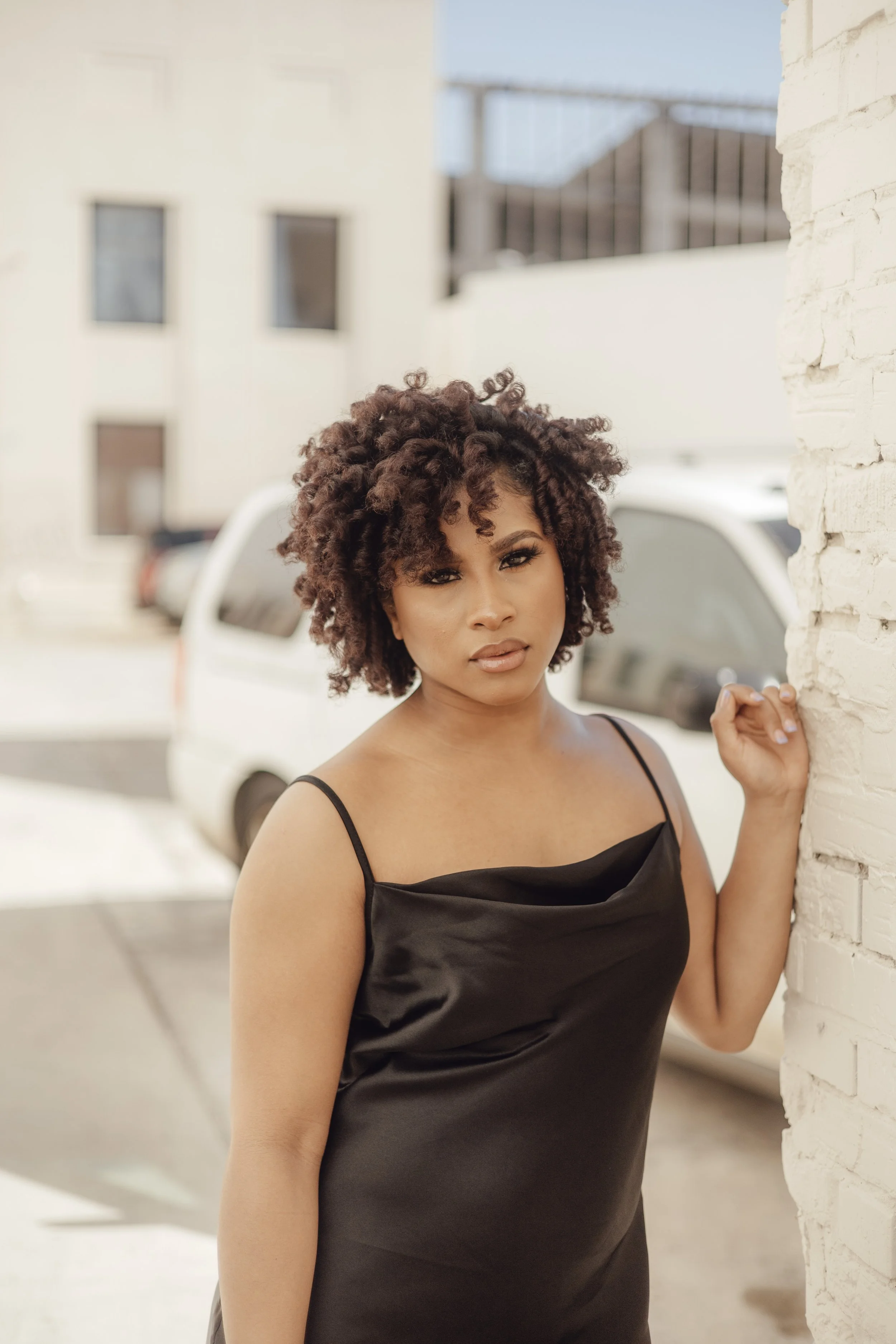 A woman with curly hair in a satin black dress standing next to a white brick wall, holding onto the wall with her right hand, with a cityscape and parked cars in the background.