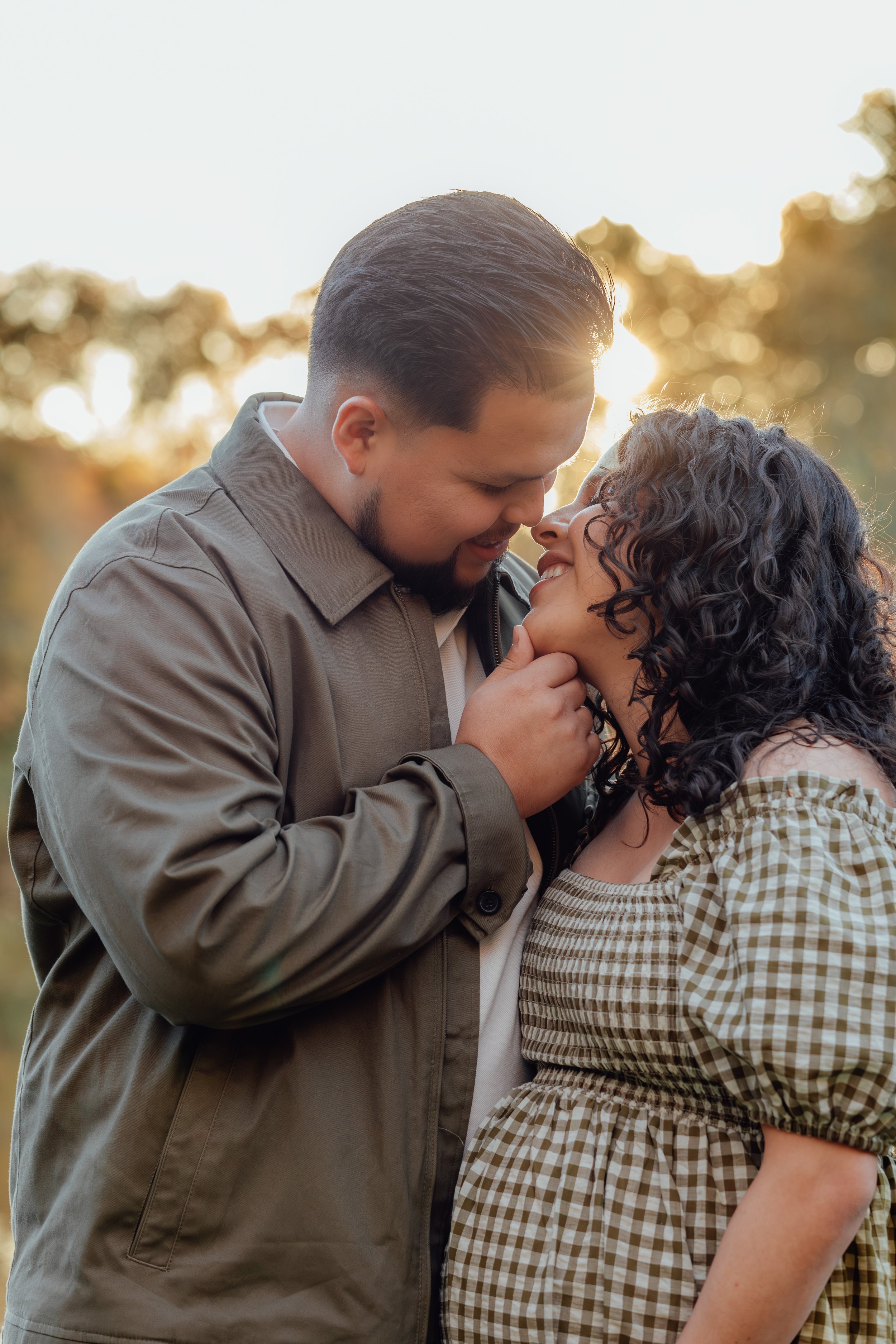 A man and woman share an intimate moment outdoors during sunset, with their foreheads touching and eyes closed, smiling while holding each other's faces.