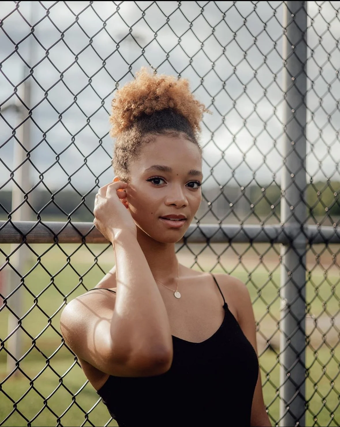 A young woman with curly, blonde-tipped hair in a high bun, standing behind a chain-link fence at a sports field during daylight, wearing a black tank top and a necklace.