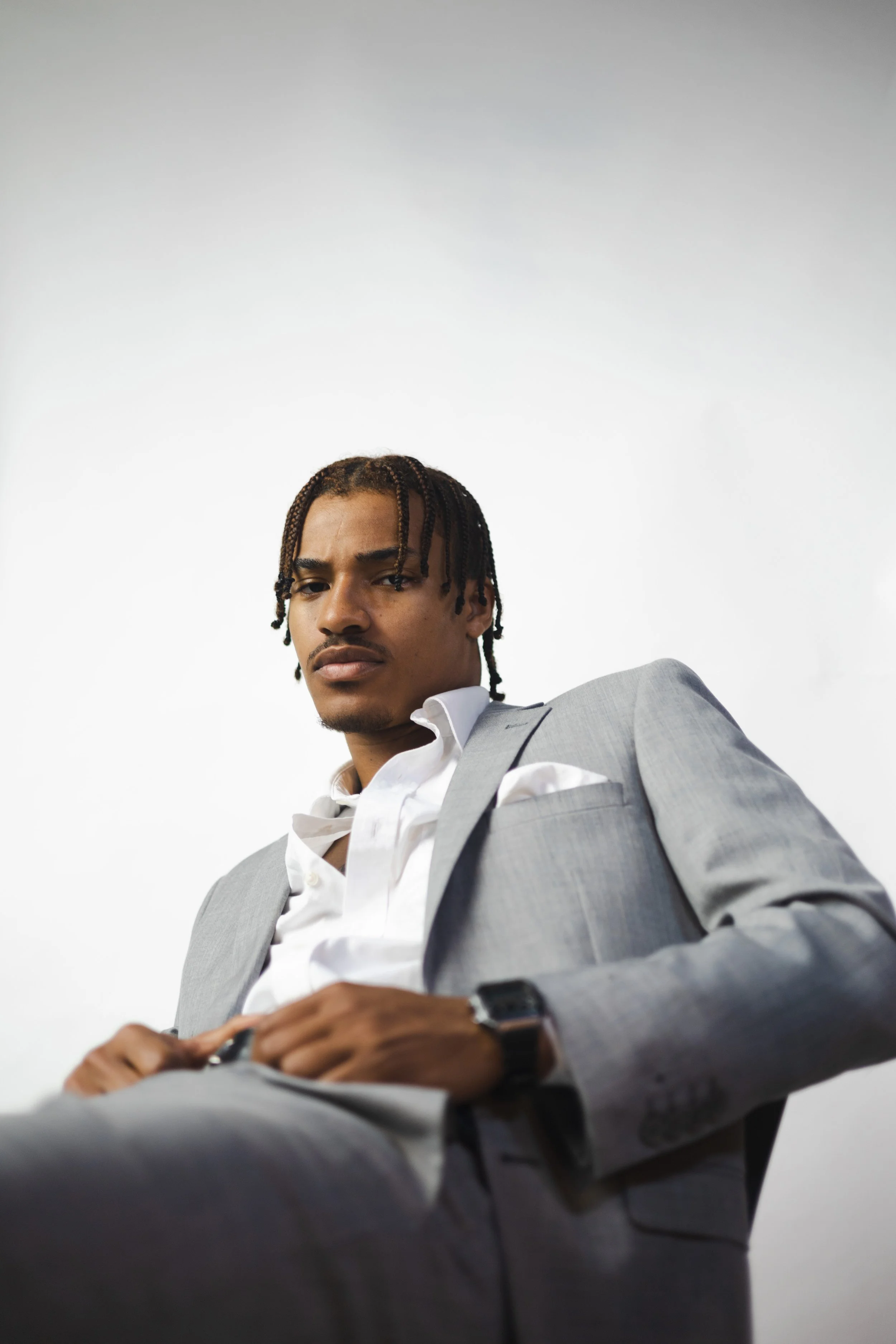 A young man with braided hair wearing a light gray suit, white shirt, and a black smartwatch on his left wrist, sitting against a plain white background.