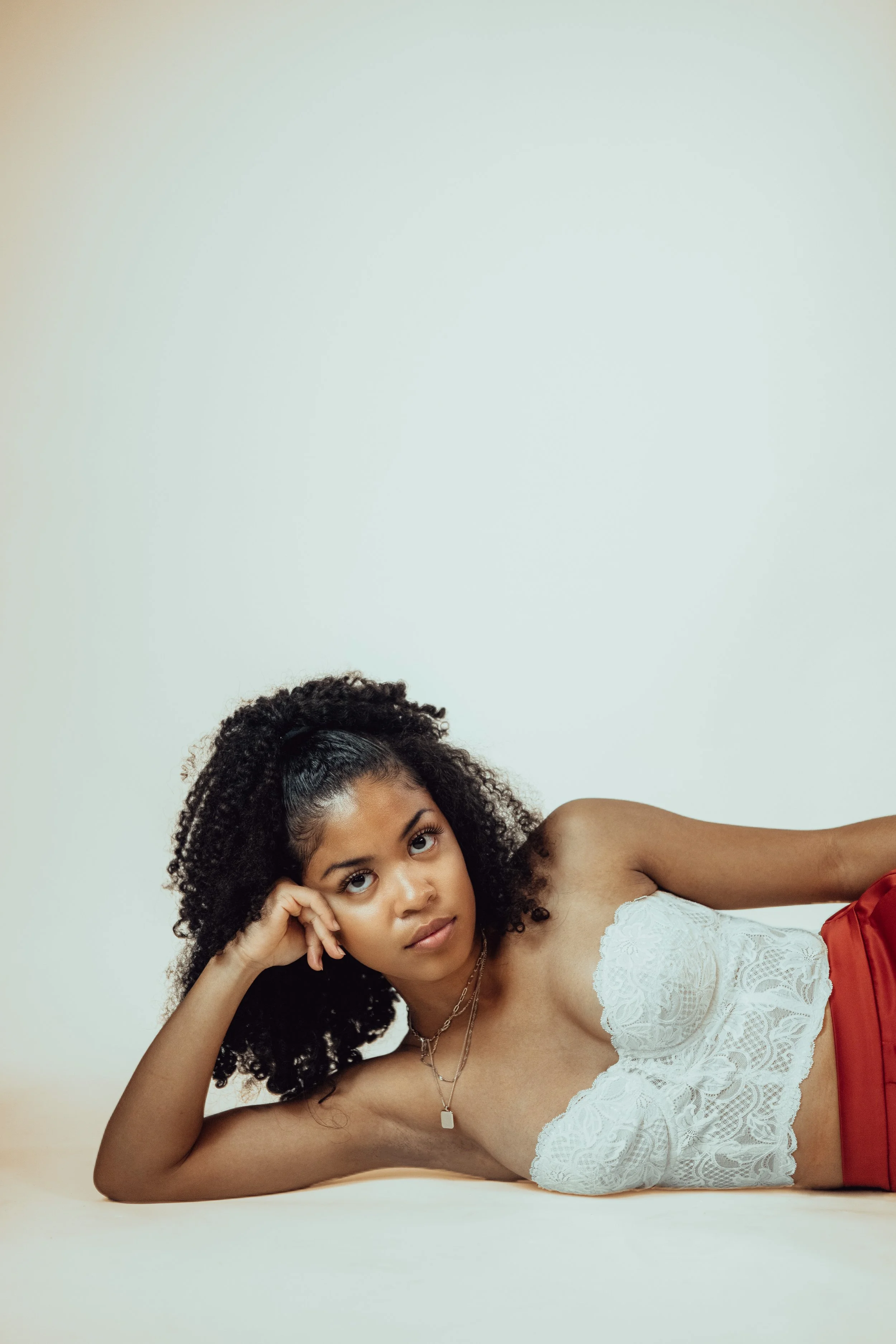 A young woman with curly hair lying on her side, wearing a white lace top and red skirt, posing against a plain light-colored background.