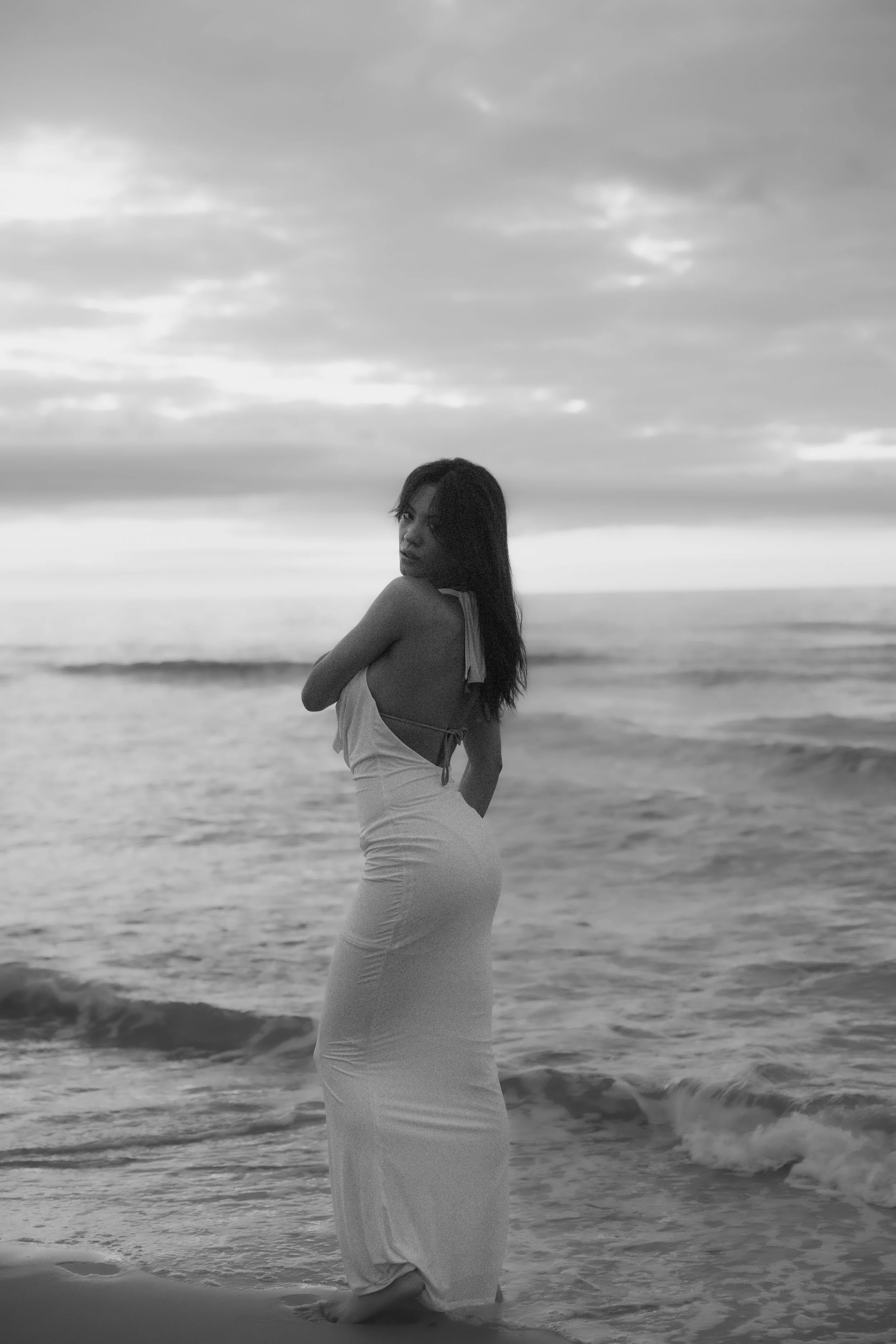 A woman in a long dress standing in shallow ocean water at sunset, looking back over her shoulder.