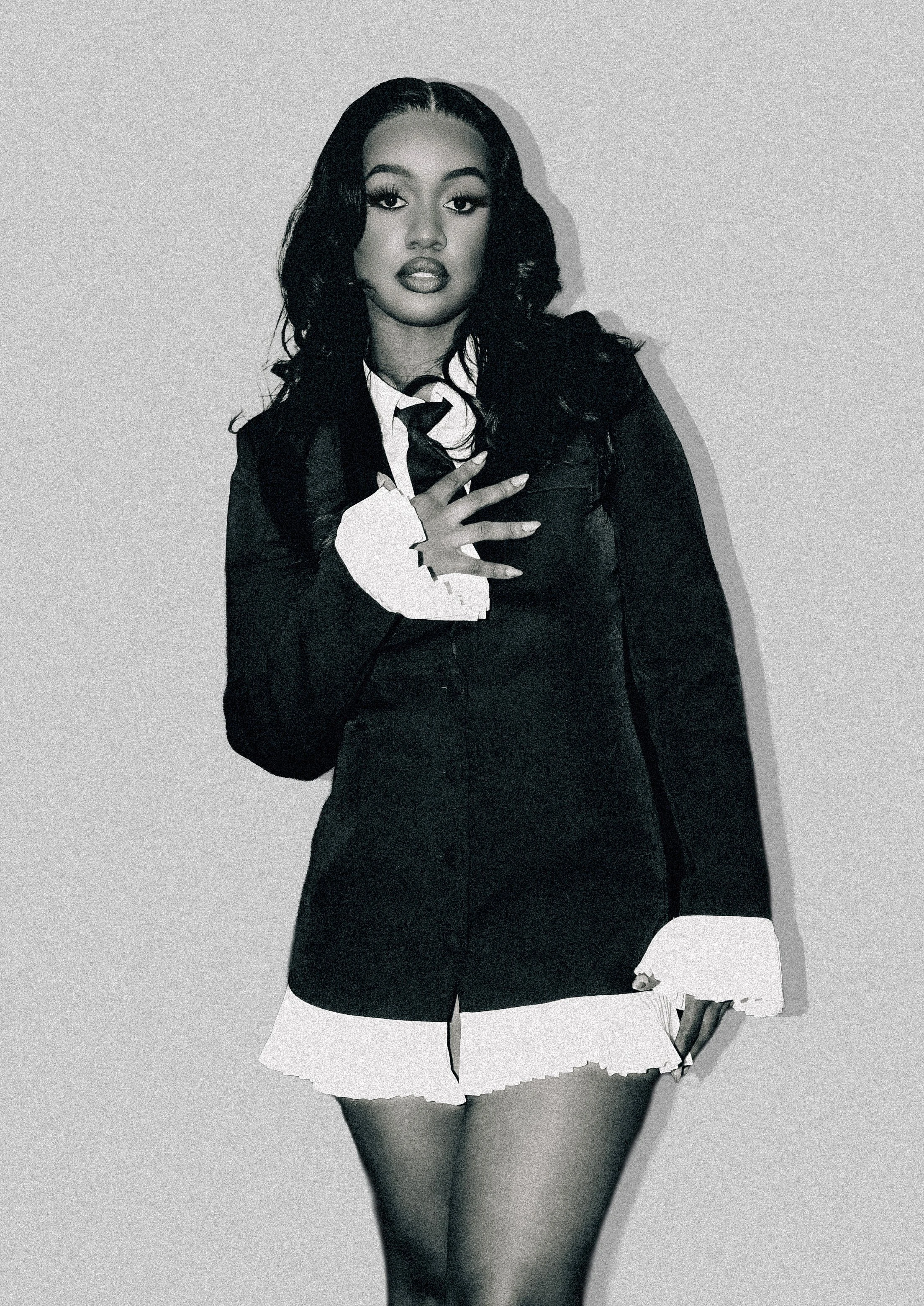 Black and white photo of a woman wearing a suit with a tie and a ruffled skirt, standing against a plain background. She is looking directly at the camera with her hand on her chest.