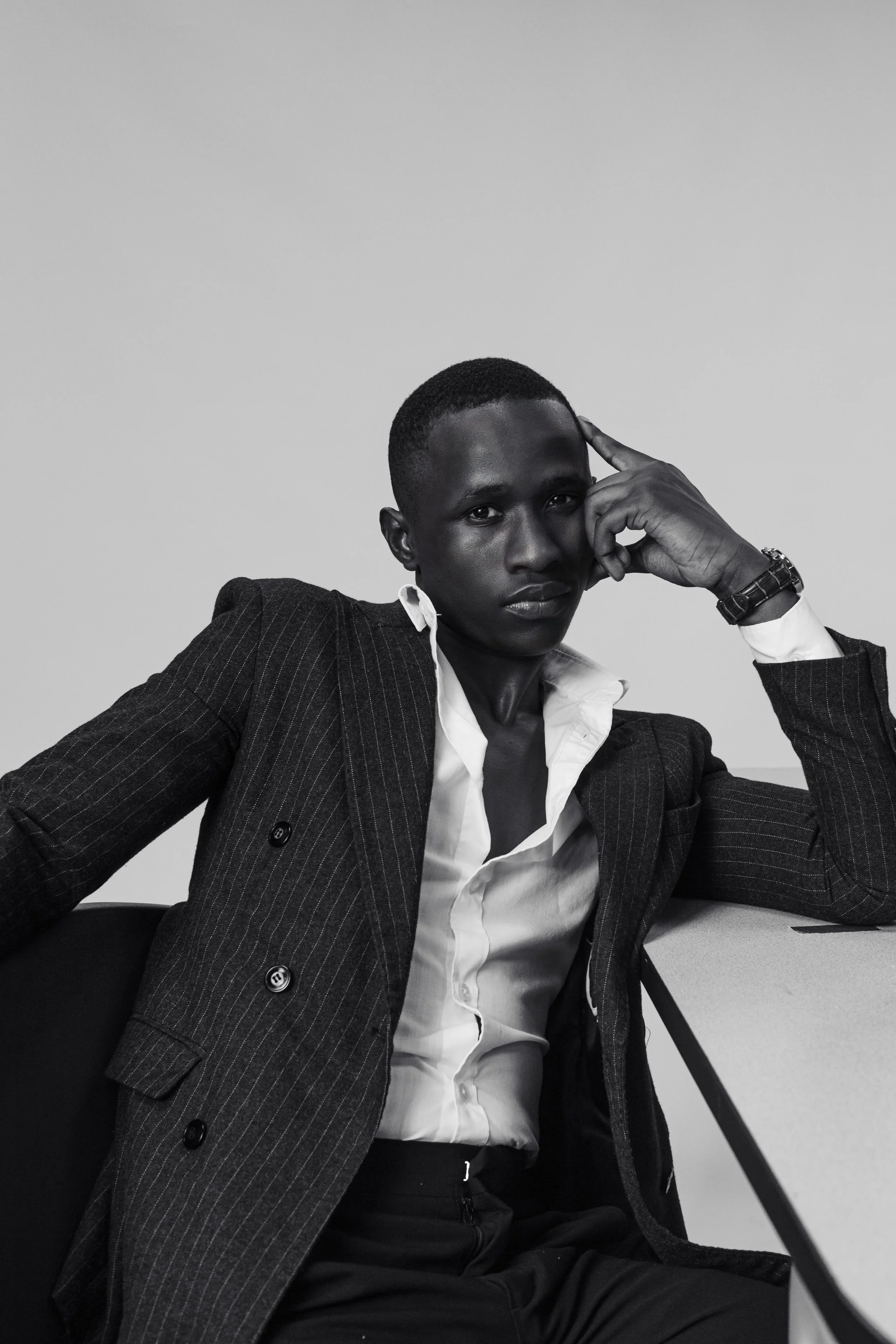 A young man in a pinstripe blazer, white shirt, and dark pants sitting at a table with a neutral expression, resting his head on his left hand, in a black-and-white photo.