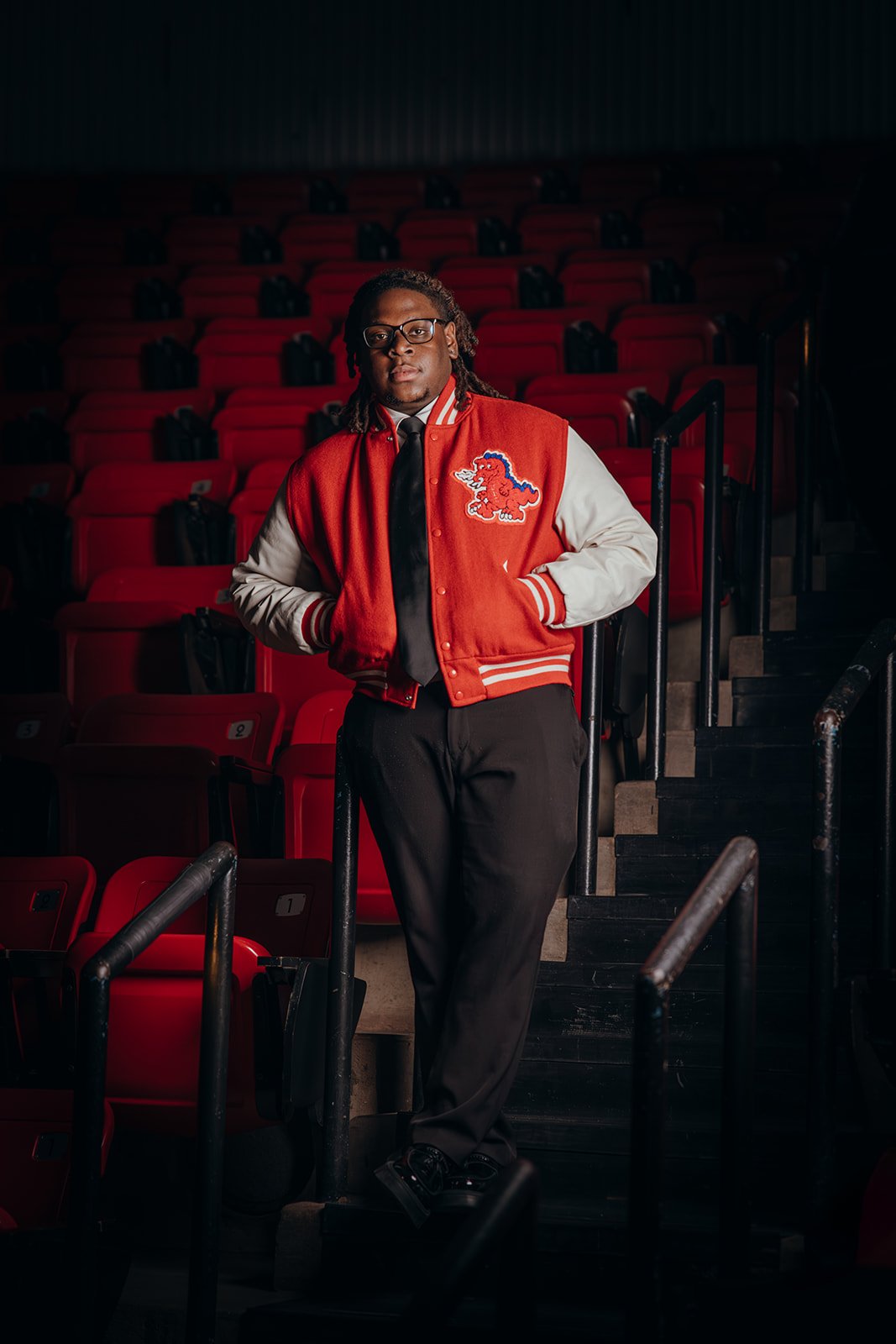 A man in a red and white varsity jacket with a lion patch, black trousers, black shoes, and glasses standing in an empty theater with red seats.