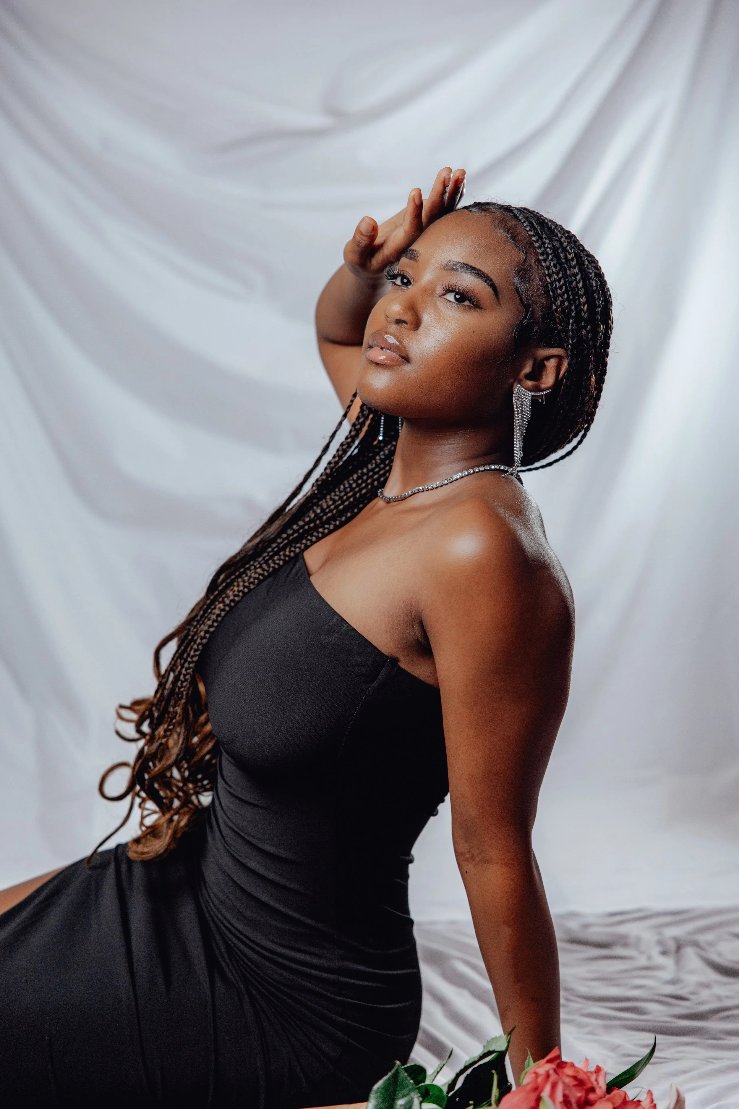 Young woman with braided hair and jewelry, wearing a black dress, posing in front of a white backdrop with flowers at the bottom, looking slightly upward with hand touching her forehead.
