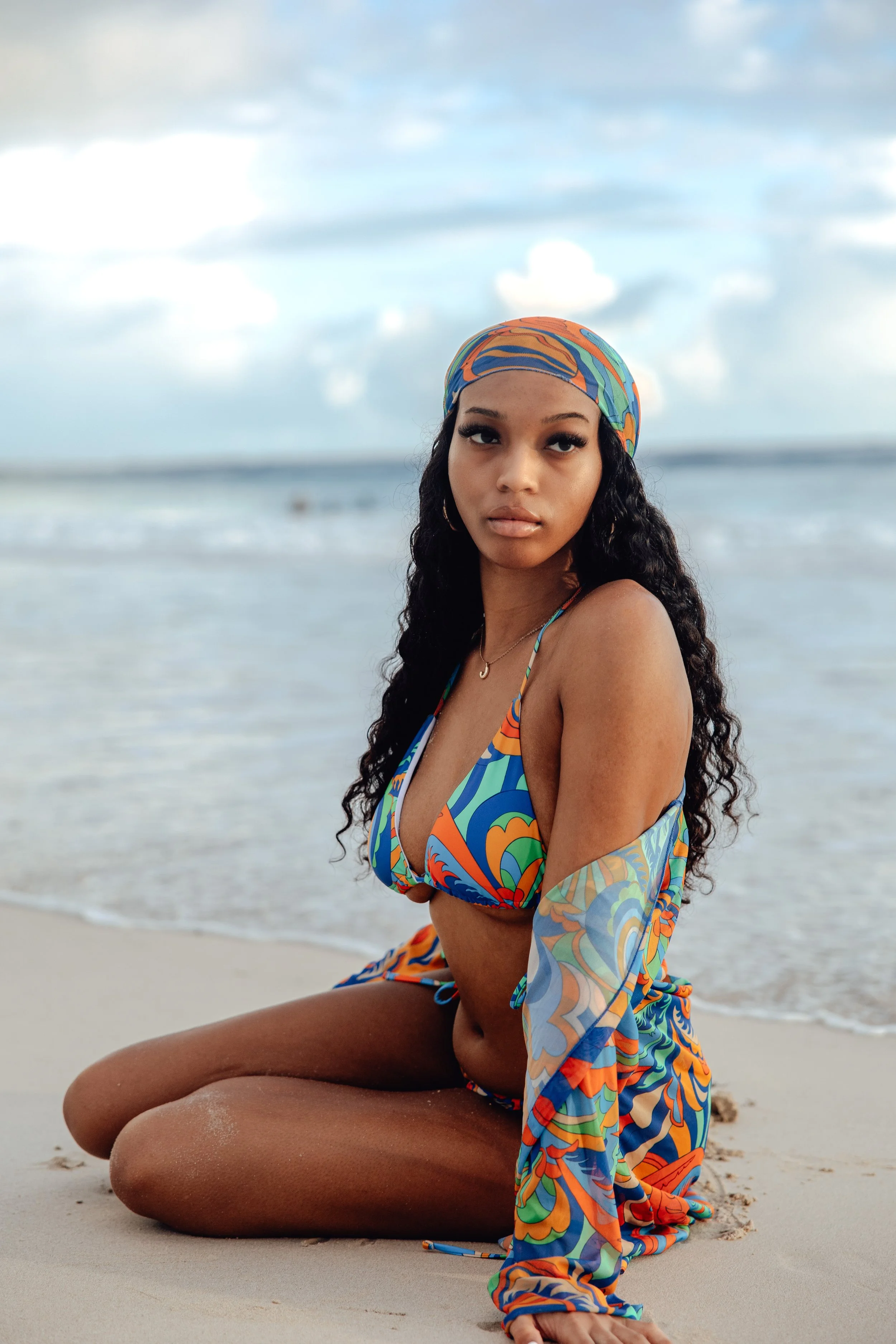 A woman with curly hair wearing a colorful bikini and matching headscarf sitting on the beach near the water, with her knee bent and looking into the camera.