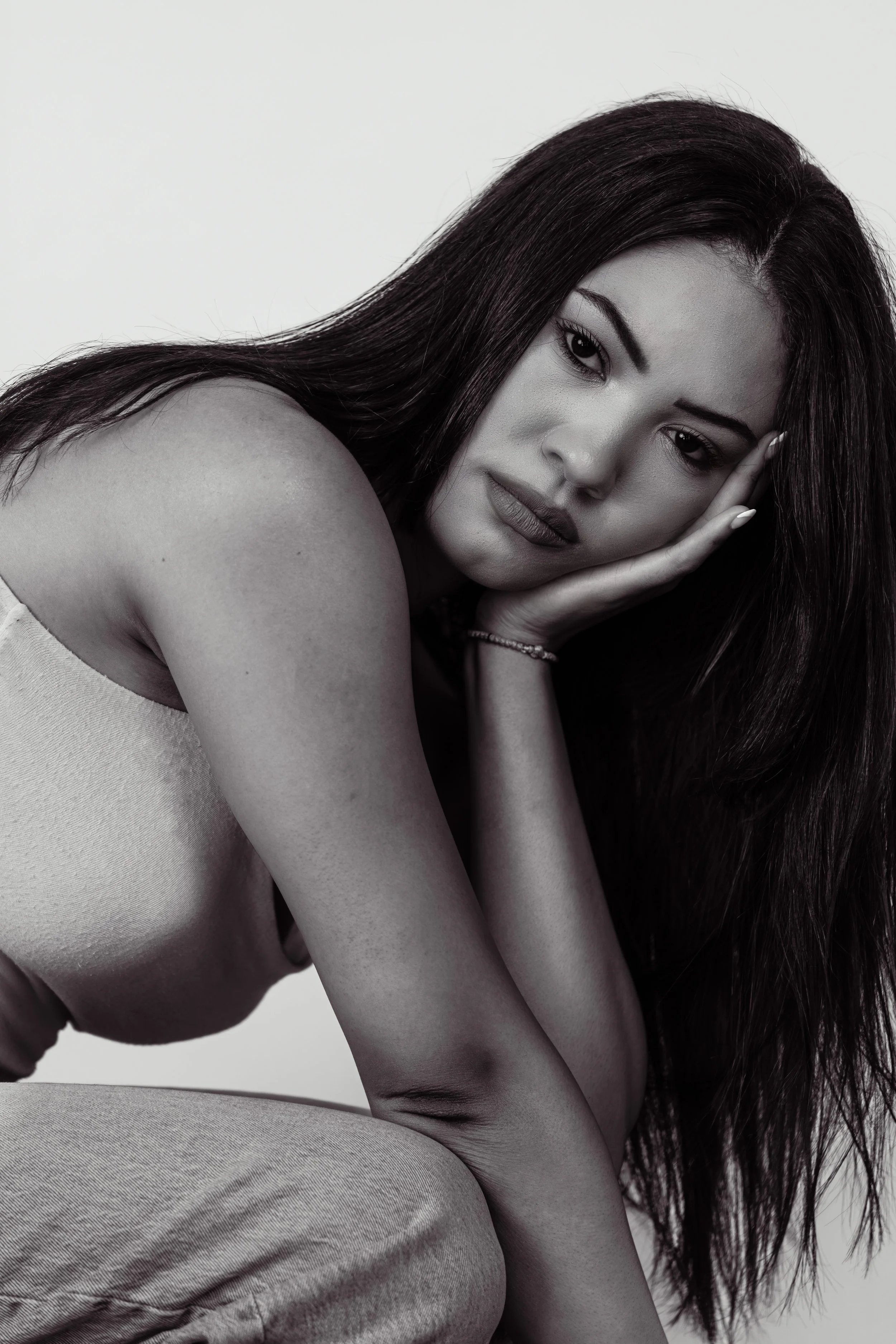 Black and white photo of a woman with dark hair, her head resting on her hand, her shoulder exposed, wearing a sleeveless top and sitting in a relaxed pose.