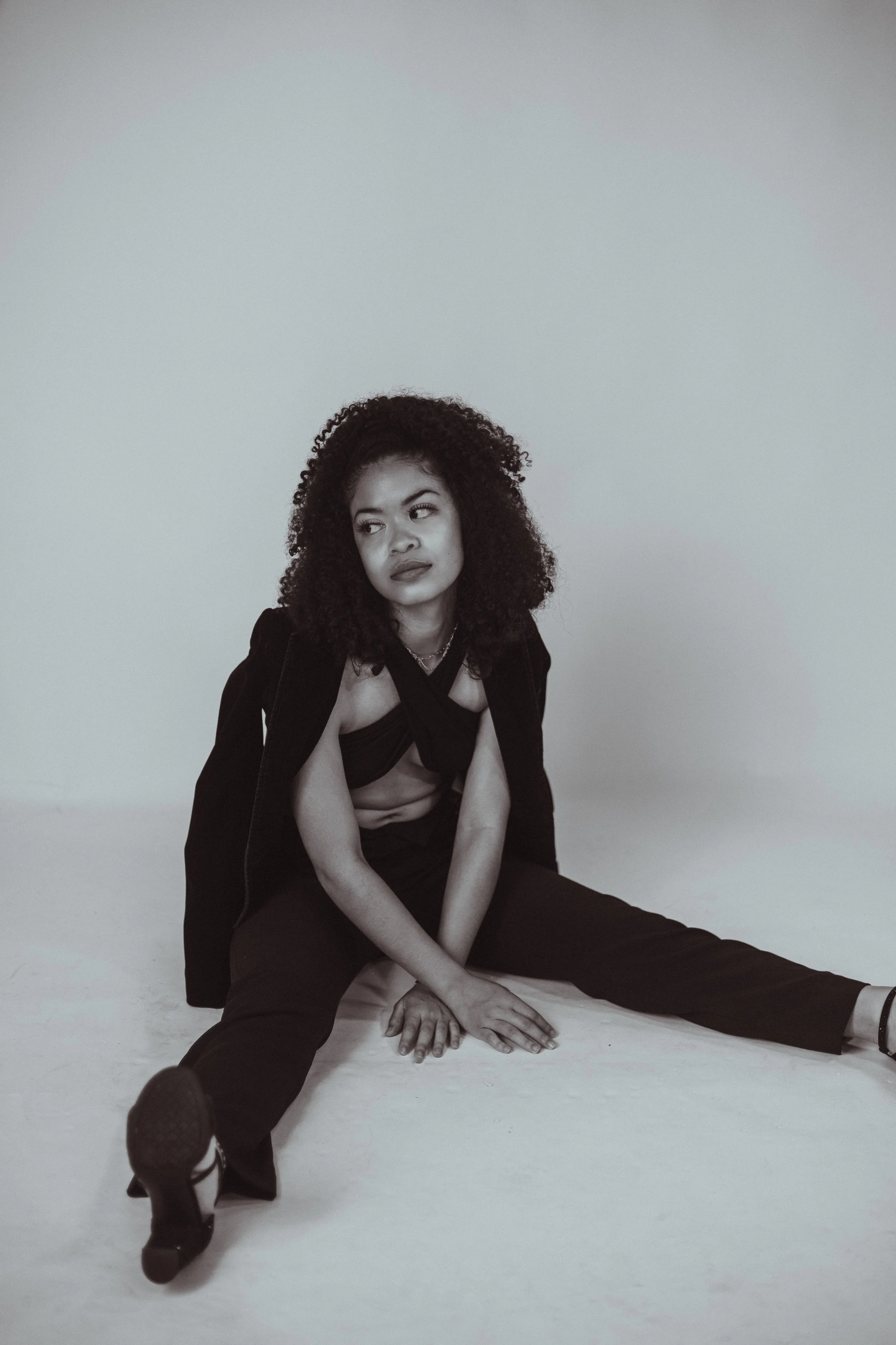 A woman with curly hair sitting on the floor in a pose, wearing a black jacket and pants, with a neutral expression, against a plain light background.