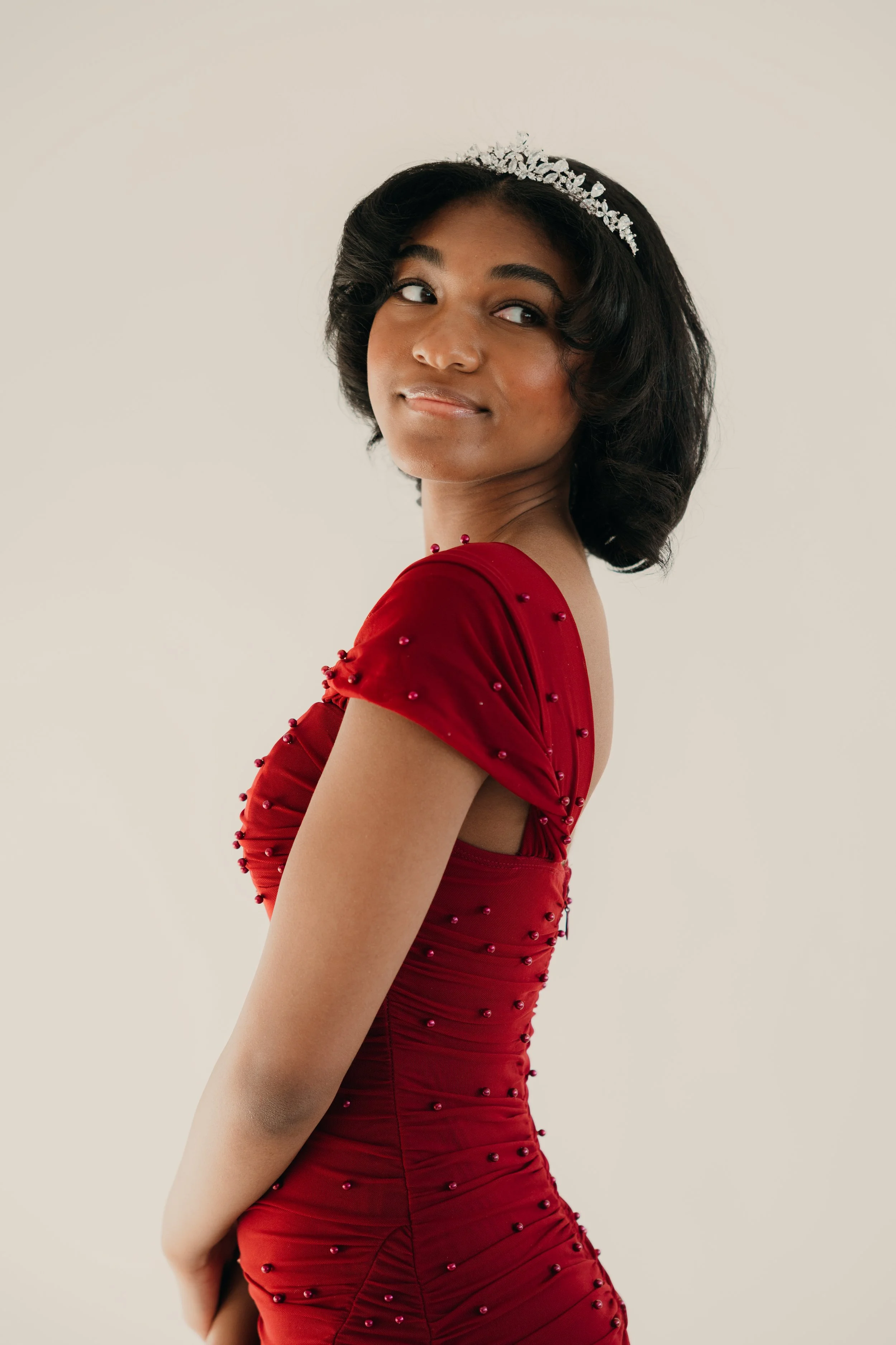 A young woman with dark curly hair wearing a silver tiara and a red dress with decorative beads, looking over her shoulder in front of a plain light-colored background.
