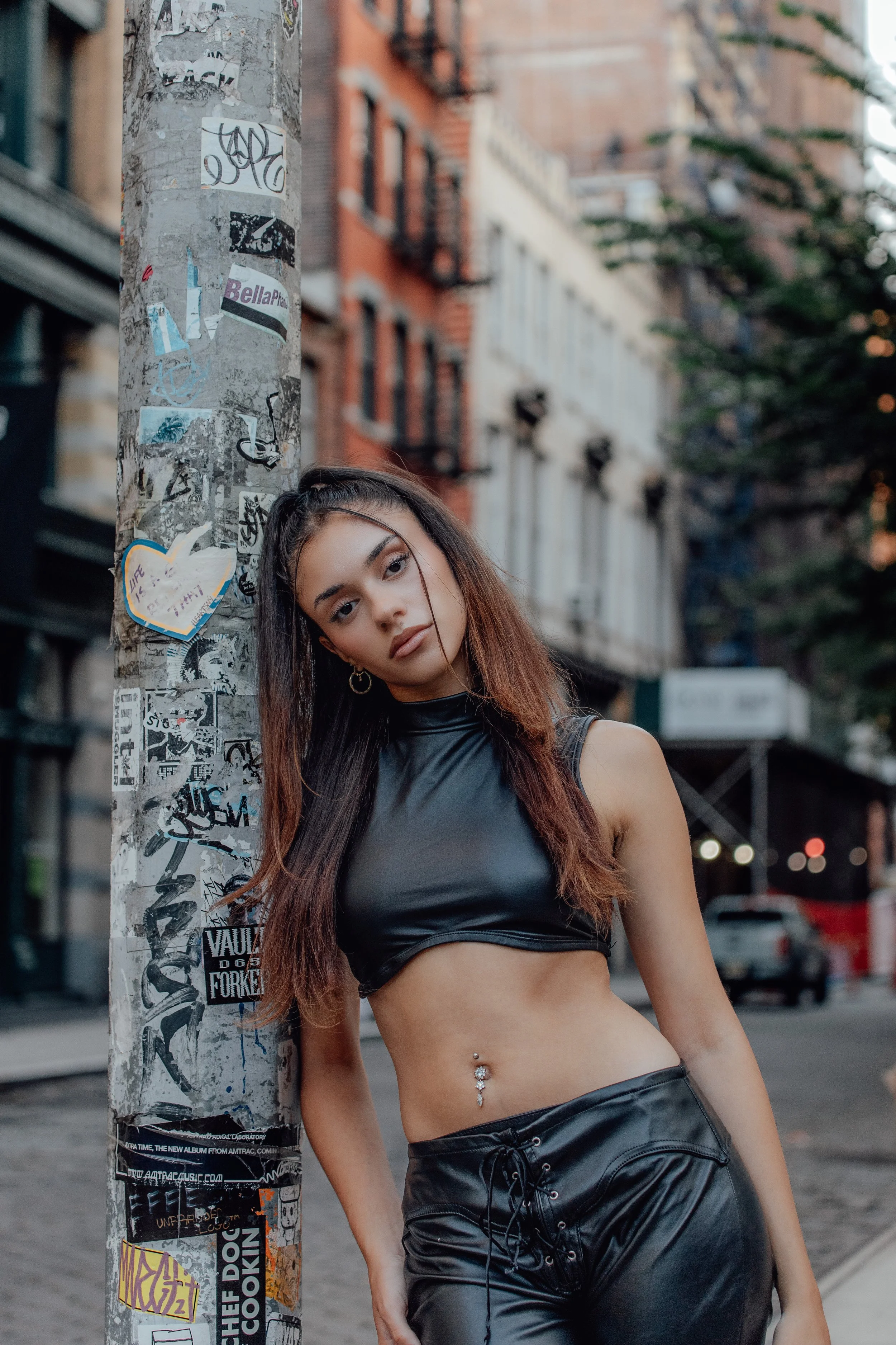 A young woman with long brown hair and belly button piercing leans against a sticker-covered utility pole on a city street during the evening. She is wearing a black sleeveless crop top and black high-waisted pants.