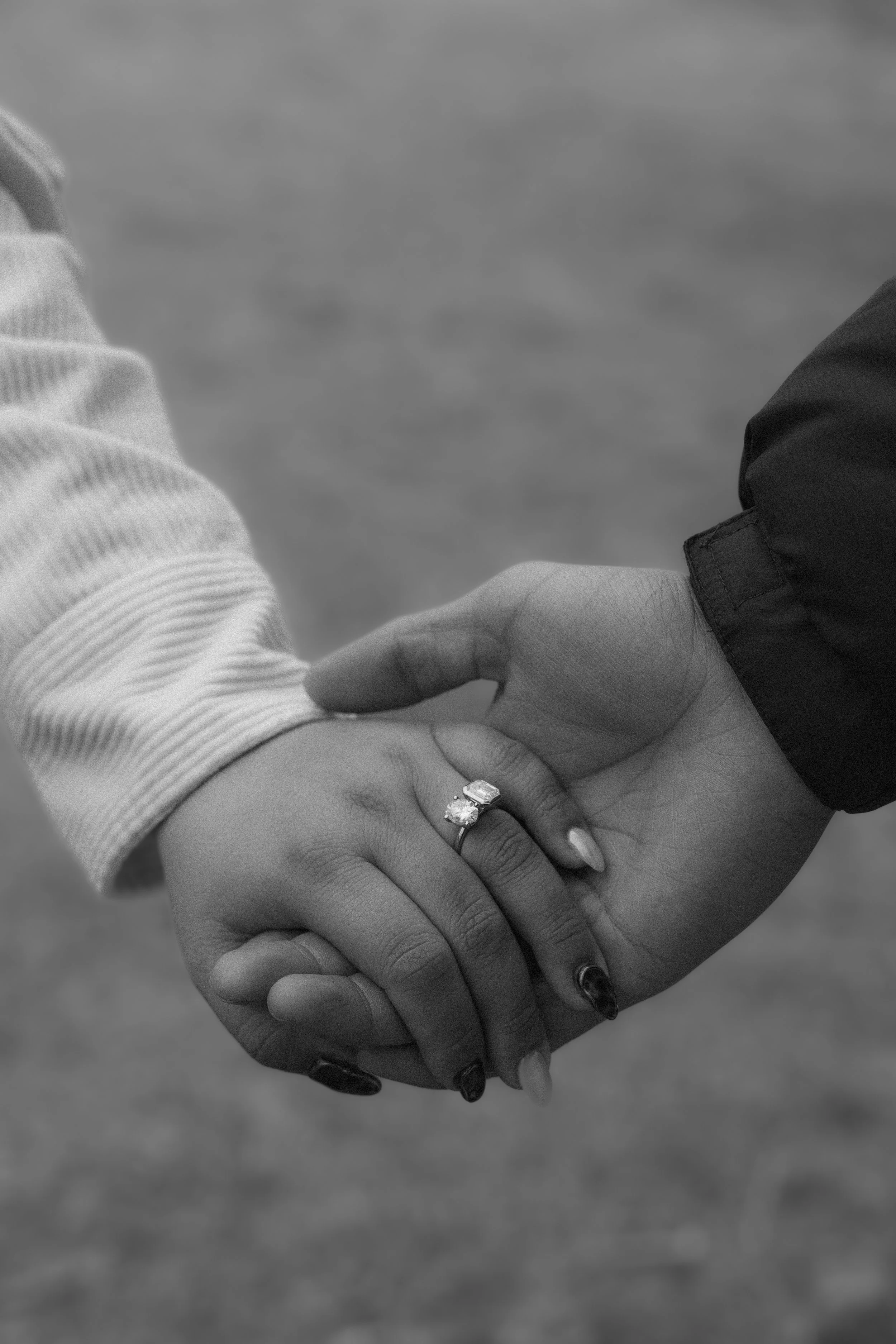 Close-up of hands holding each other, one hand with an engagement ring and a wedding band, both wearing long-sleeve shirts.