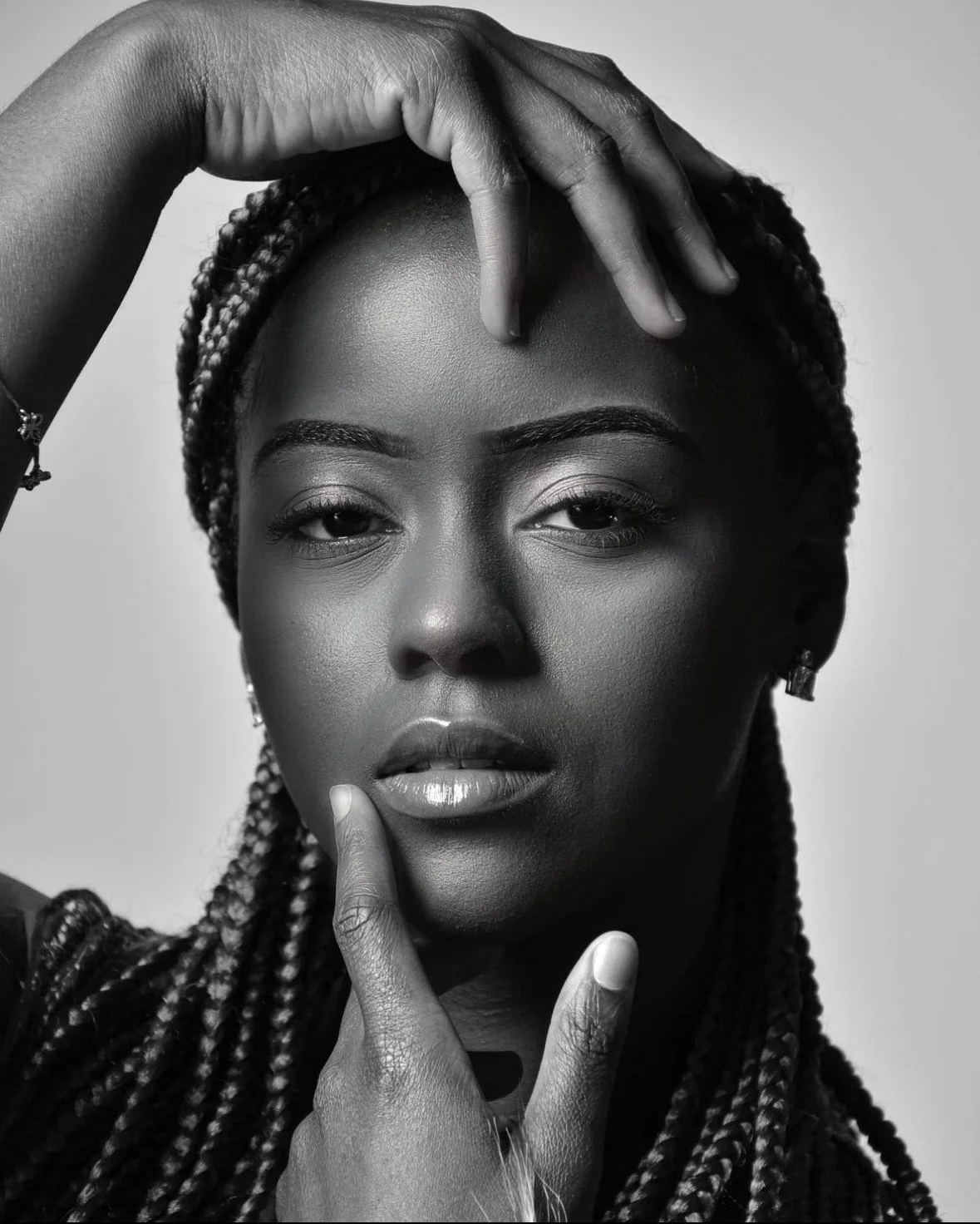 A black and white portrait of a woman with braided hair, holding her hand above her forehead and her finger near her lips, with a serious expression.
