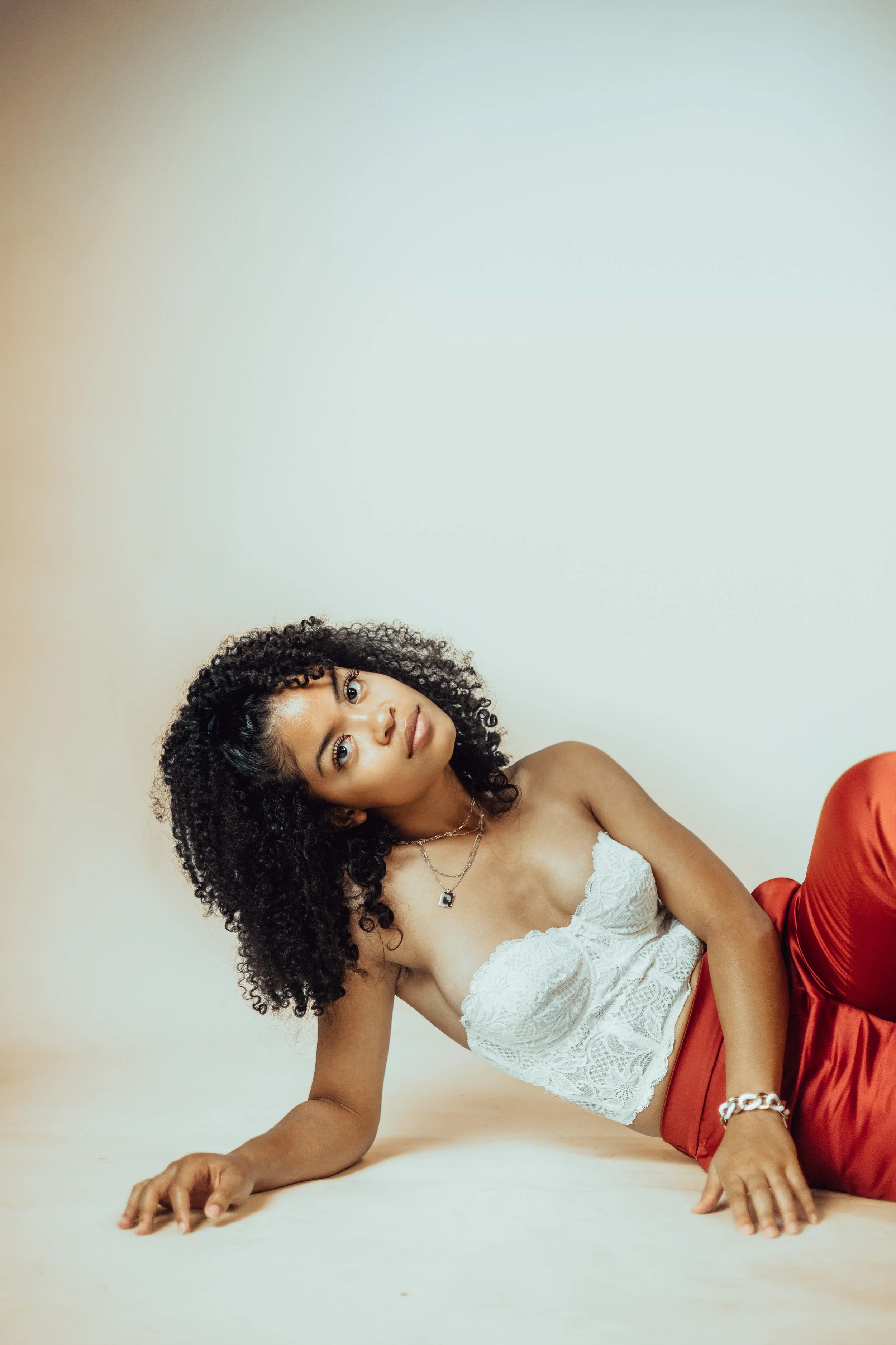 A woman with curly black hair, wearing a white lace crop top and red satin pants, lying on the floor with a neutral expression in front of a plain light-colored background.