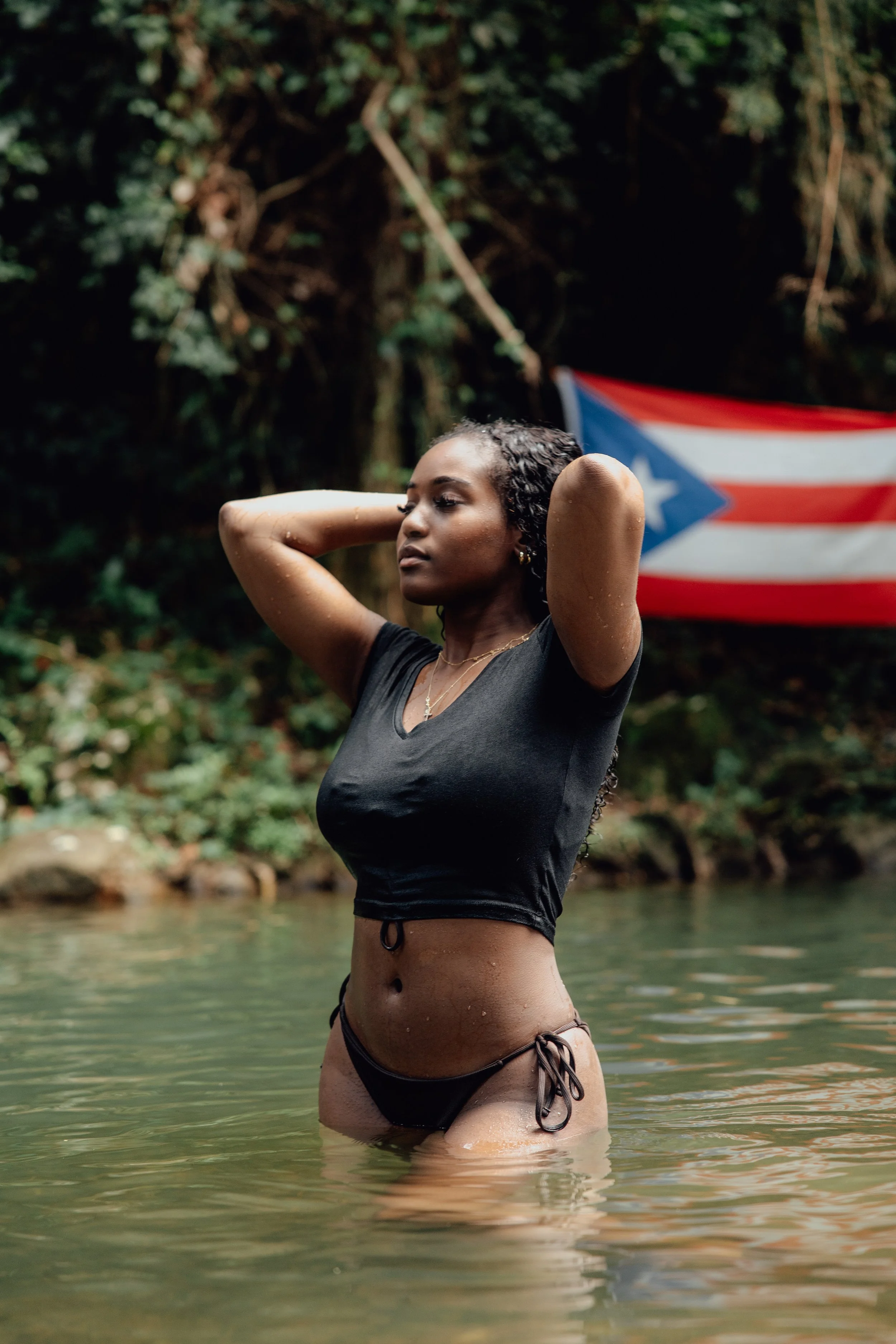 A young woman with curly hair in a black crop top and bikini bottoms stands in a shallow river, with her hands behind her head and a Puerto Rican flag waving in the background. The setting appears to be a lush, green outdoor area.