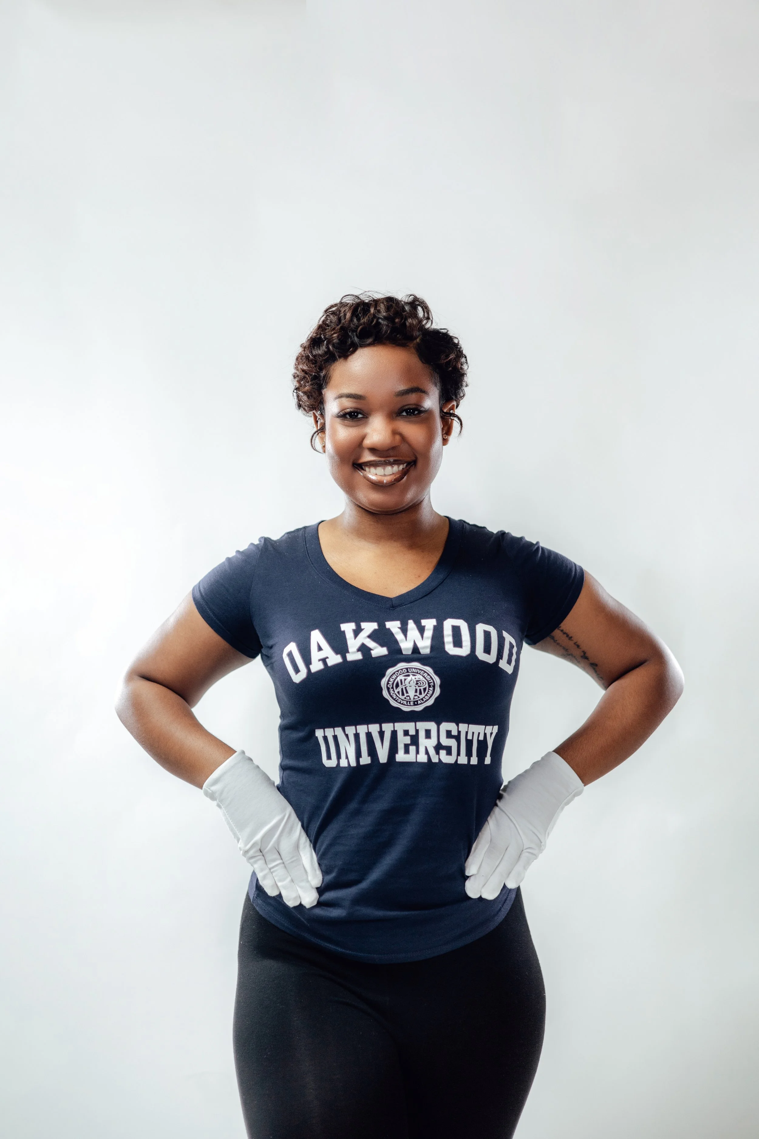 A woman with short curly hair smiling, wearing a navy t-shirt with "Oakwood University" written on it, gray gloves, black leggings, standing with hands on hips against a plain white wall.