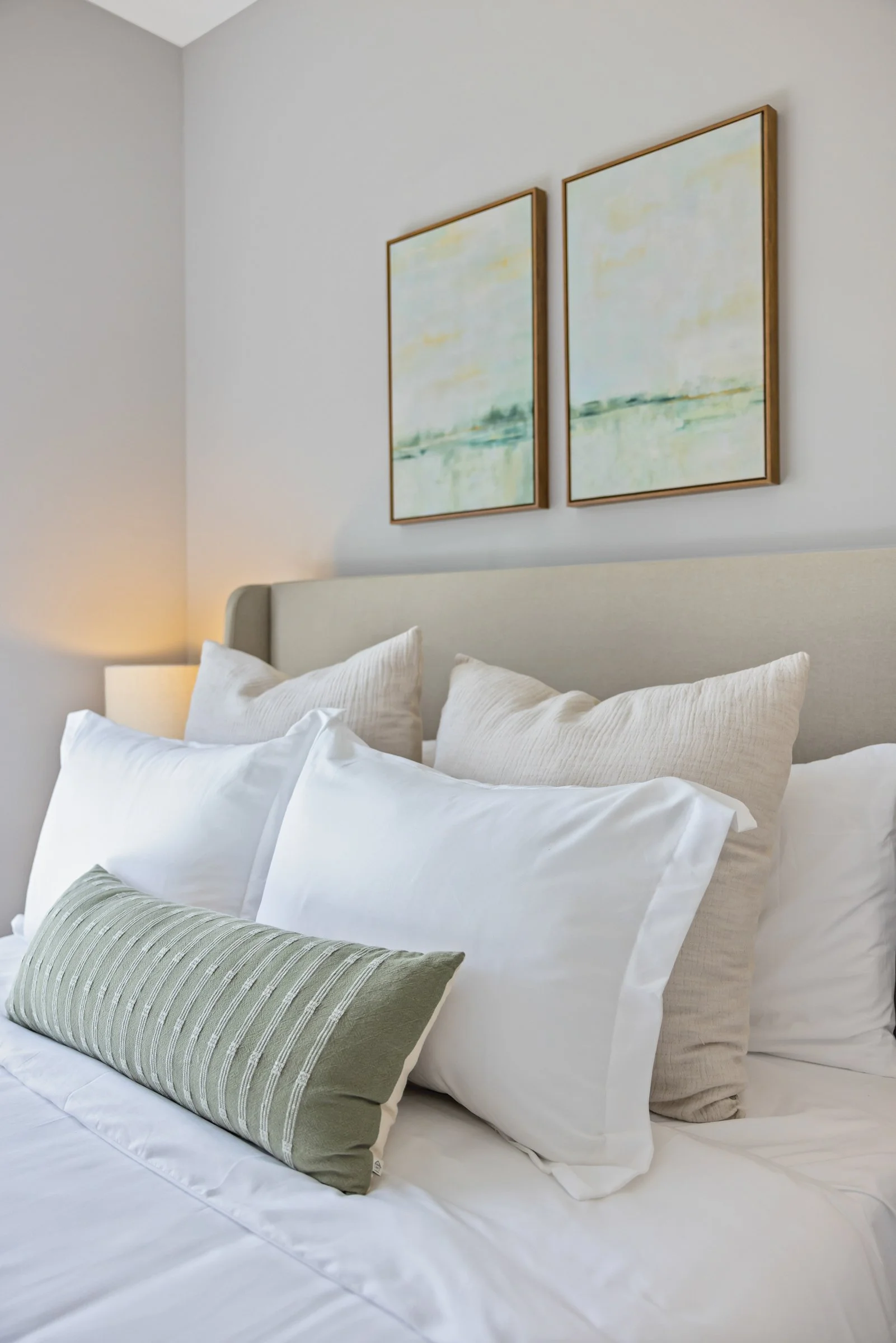 Image of a bed with white bedding and a green throw pillow with two paintings above the bed. The bed is set against ight grey walls and a lamp.