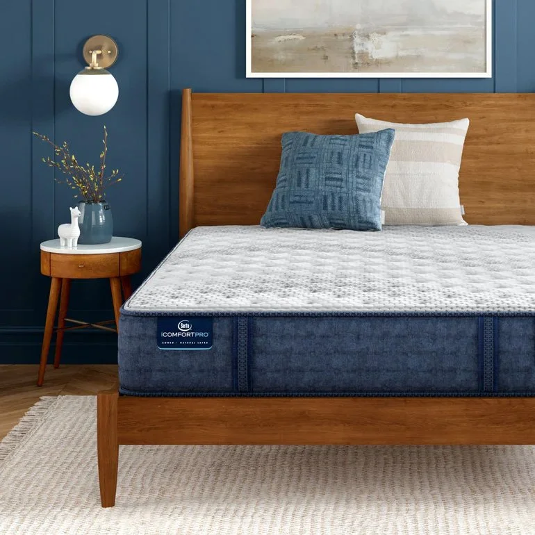 A photo of a wooden bed with a bare mattress and two throw pillows. The bed is in front of a blue wall, next to a wooden side table, and is on top of a neutral carpet.