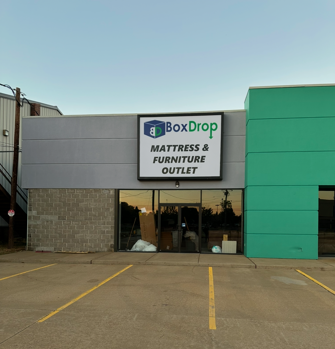 Photo of the Box Drop Mattress and Furniture outlet building and parking lot in Alexandria, LA. The building is brick, grey, and green with glass doors.