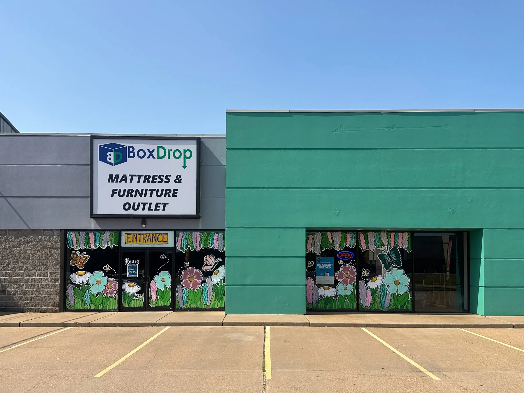 View of a store front selling mattresses and furniture, with a sign that reads 'BoxDrop Mattress & Furniture Outlet,' and decorative door with floral and butterfly designs, and an entrance sign with colorful flowers.
