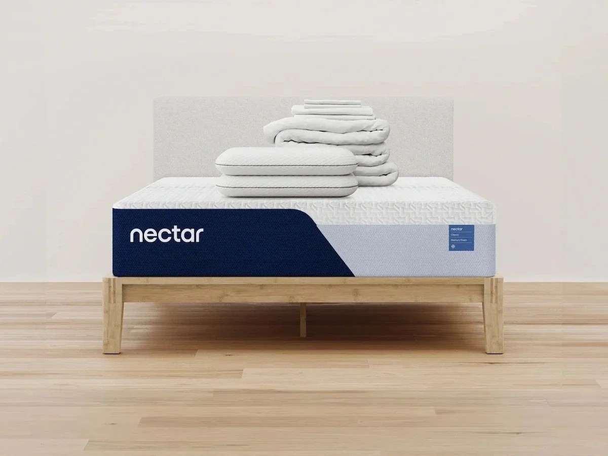A wooden bed frame and a mattress with the logo "nectar". There are folded mattress toppers on top, and the room is white with light wooden flooring.