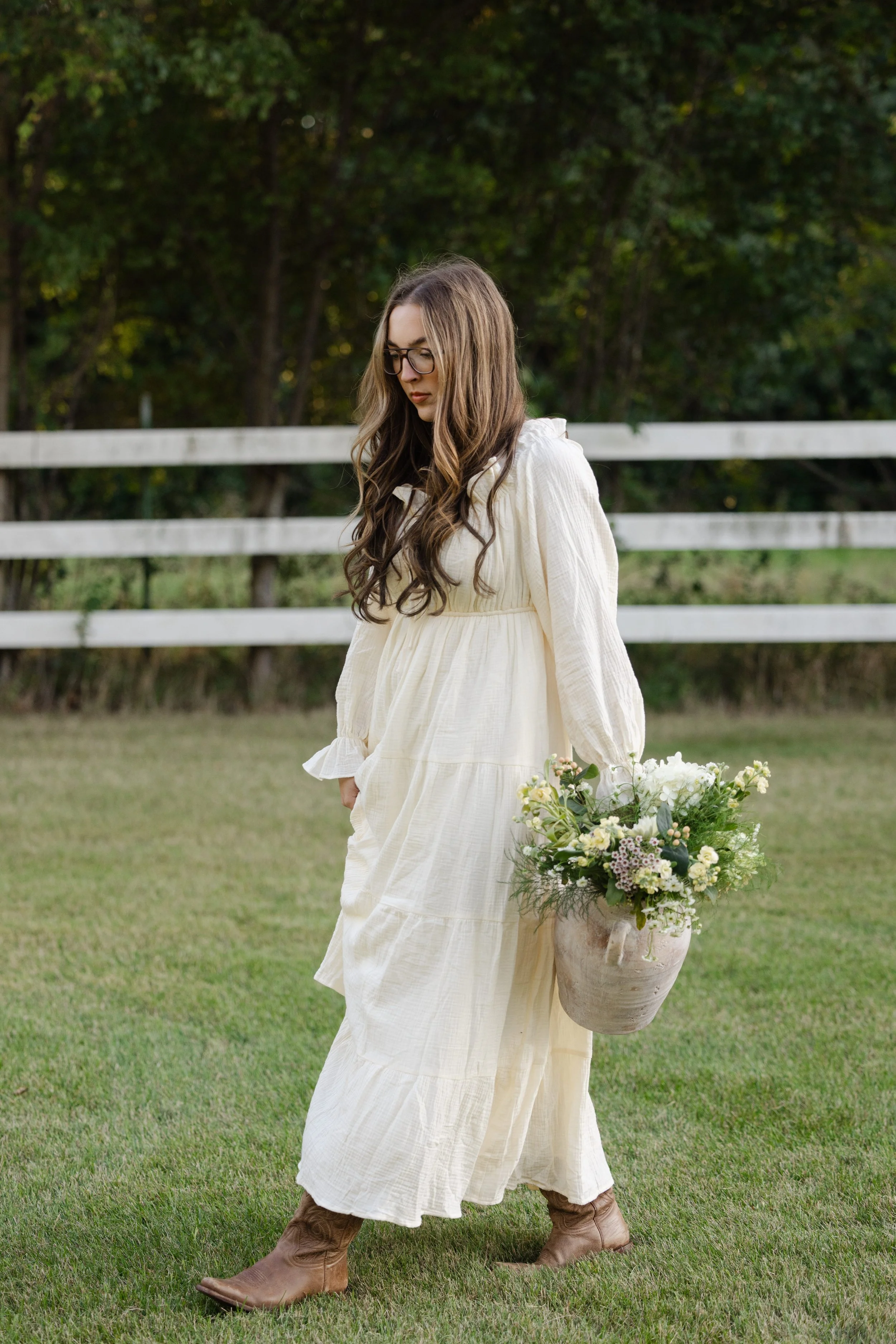 Woman wearing glasses and a white dress walks across a grassy field carrying a large bouquet of flowers.