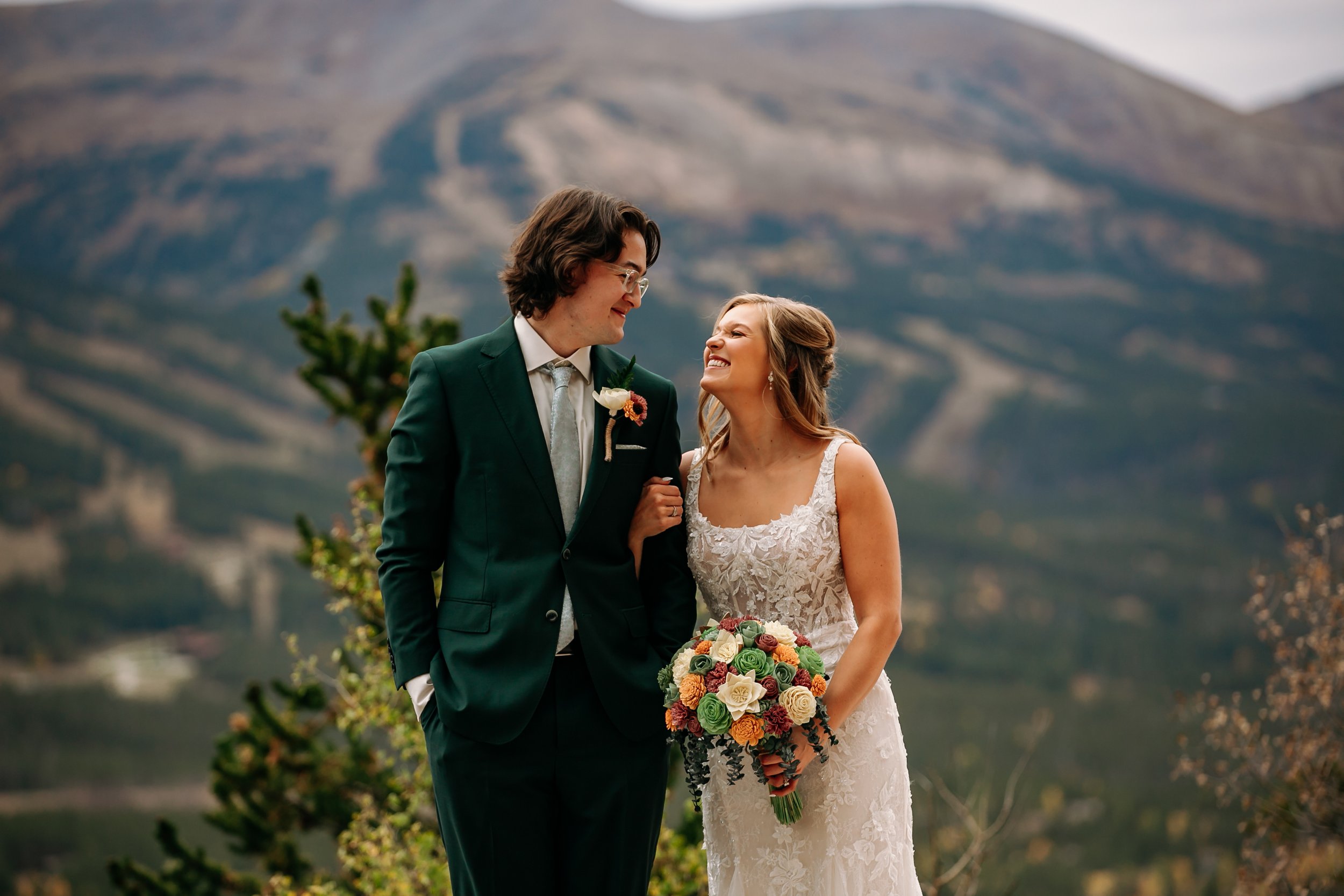 Smiling newlywed couple posing outdoors in wedding attire with a colorful bouquet and mountain landscape backdrop