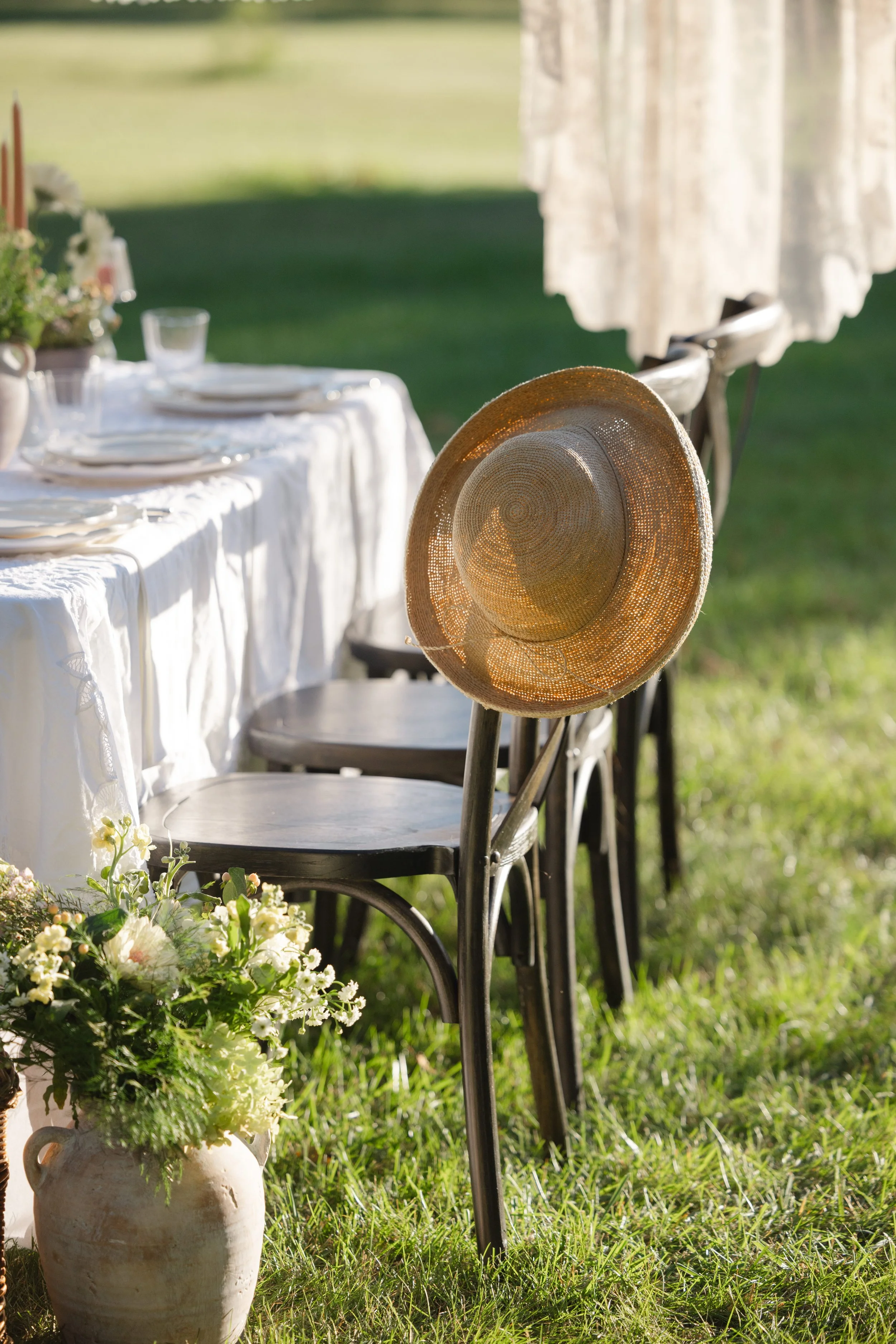 Elegant outdoor wedding table setting with white linens, floral arrangements, plates, glassware, and a straw hat on a chair