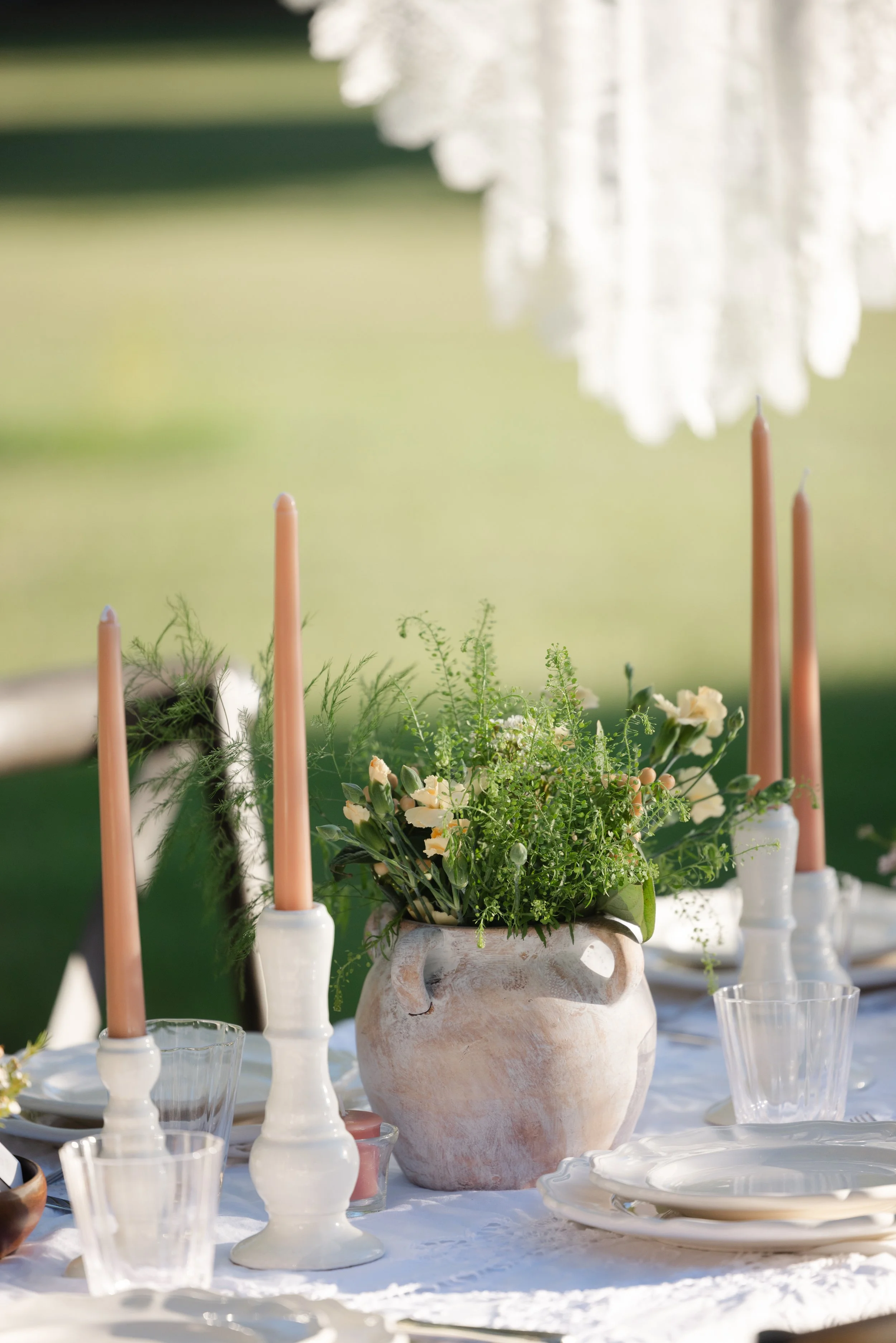Elegant outdoor table setting with peach taper candles, white plates, glassware, and a lush floral centerpiece in a rustic ceramic vase