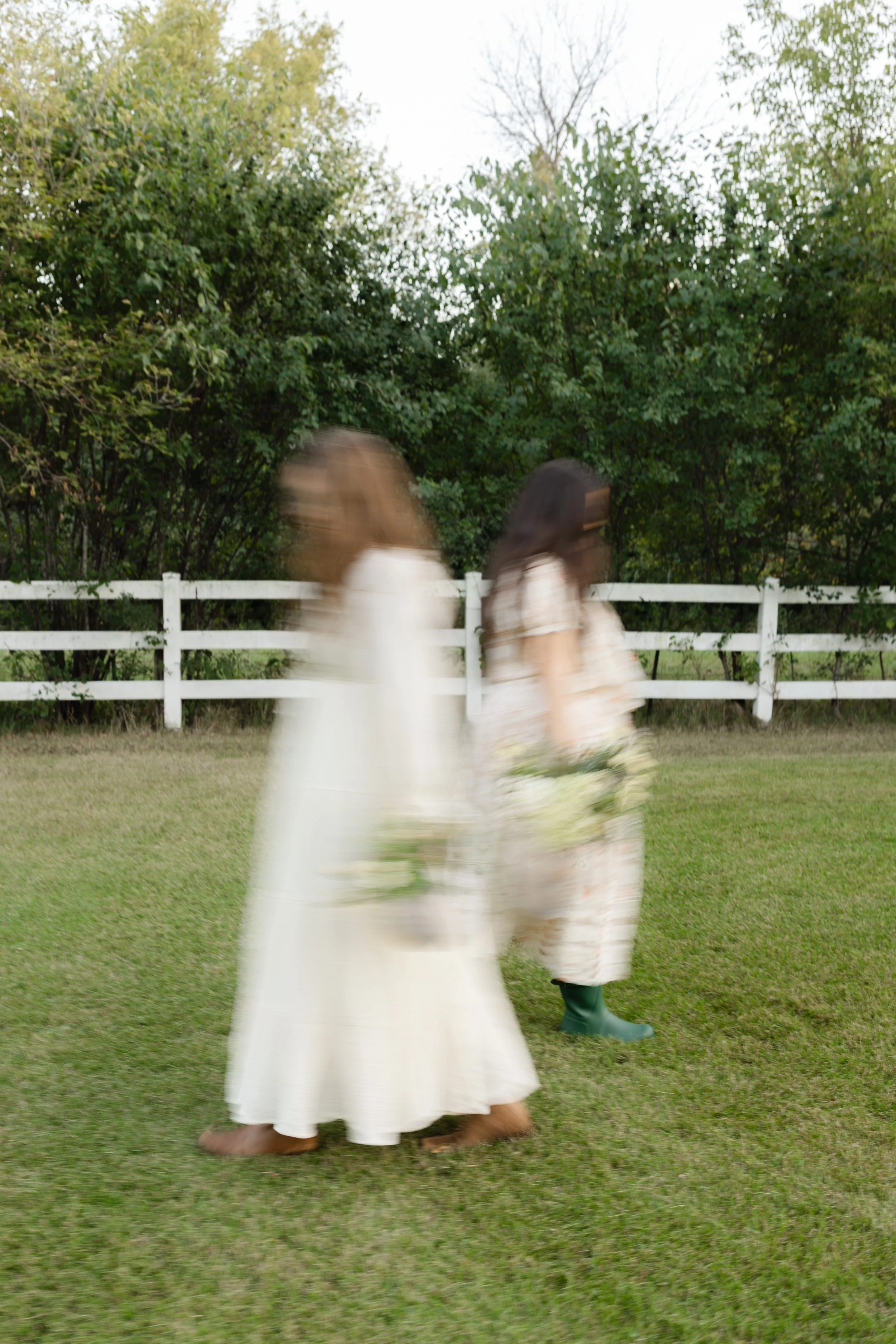 Motion-blurred people walking across a grassy yard in front of a white fence and dense trees
