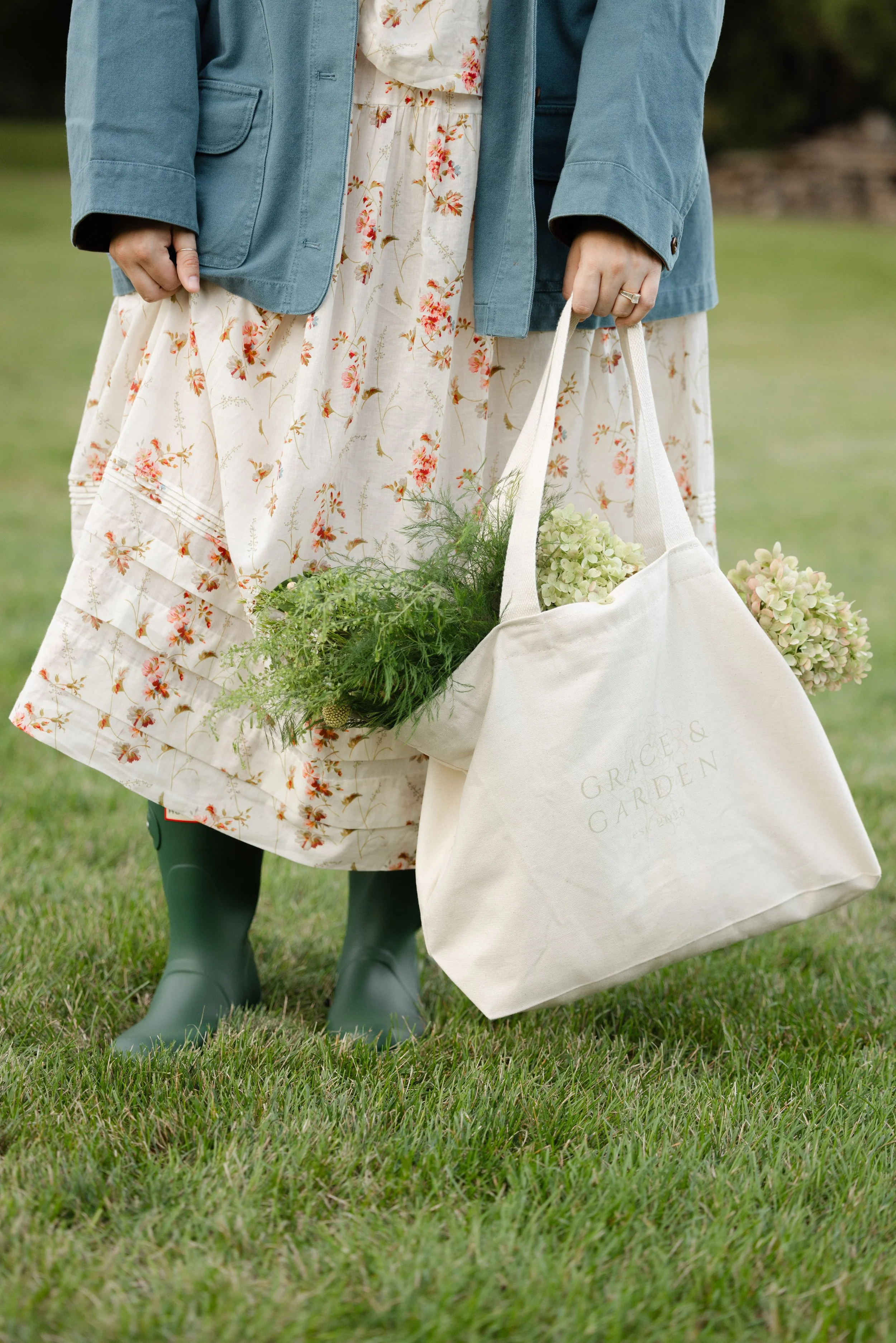 Woman in a blue jacket and floral dress carrying a canvas tote filled with flowers outdoors