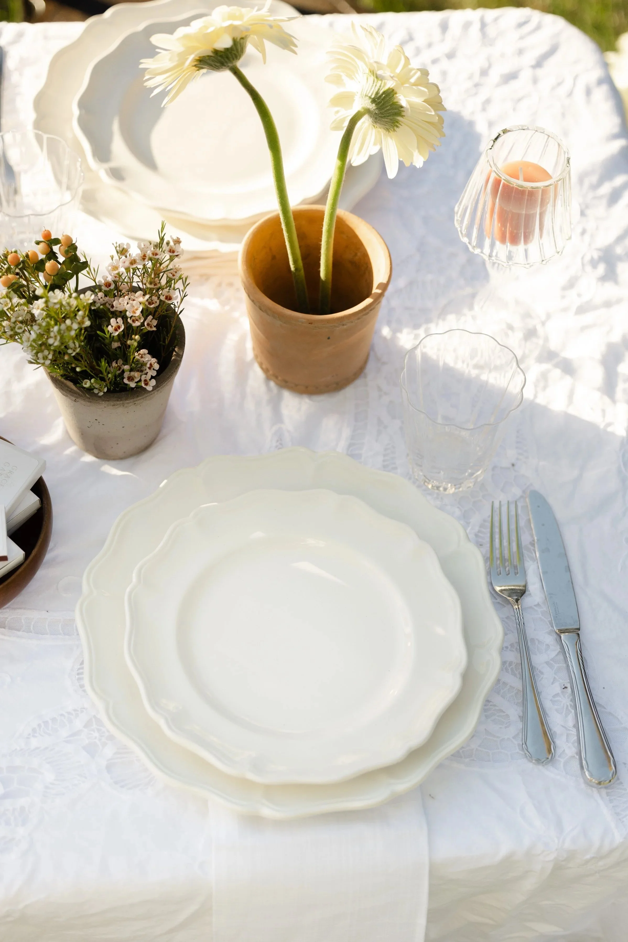 Elegant outdoor table setting with white plates, silverware, potted white daisies, flowers, and a candle on a white lace tablecloth