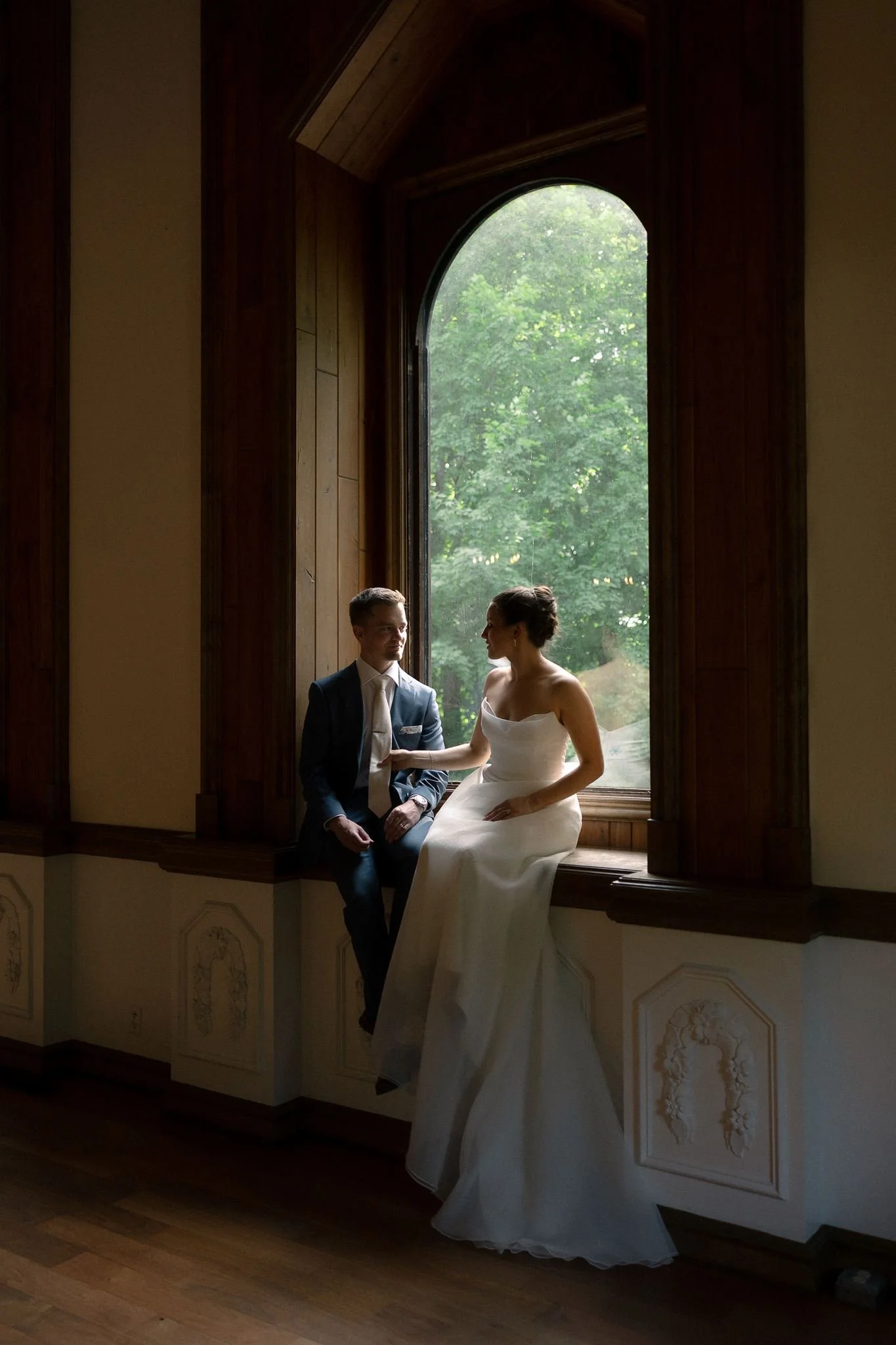 Bride and groom sitting together on a window ledge in an elegant wood-paneled room