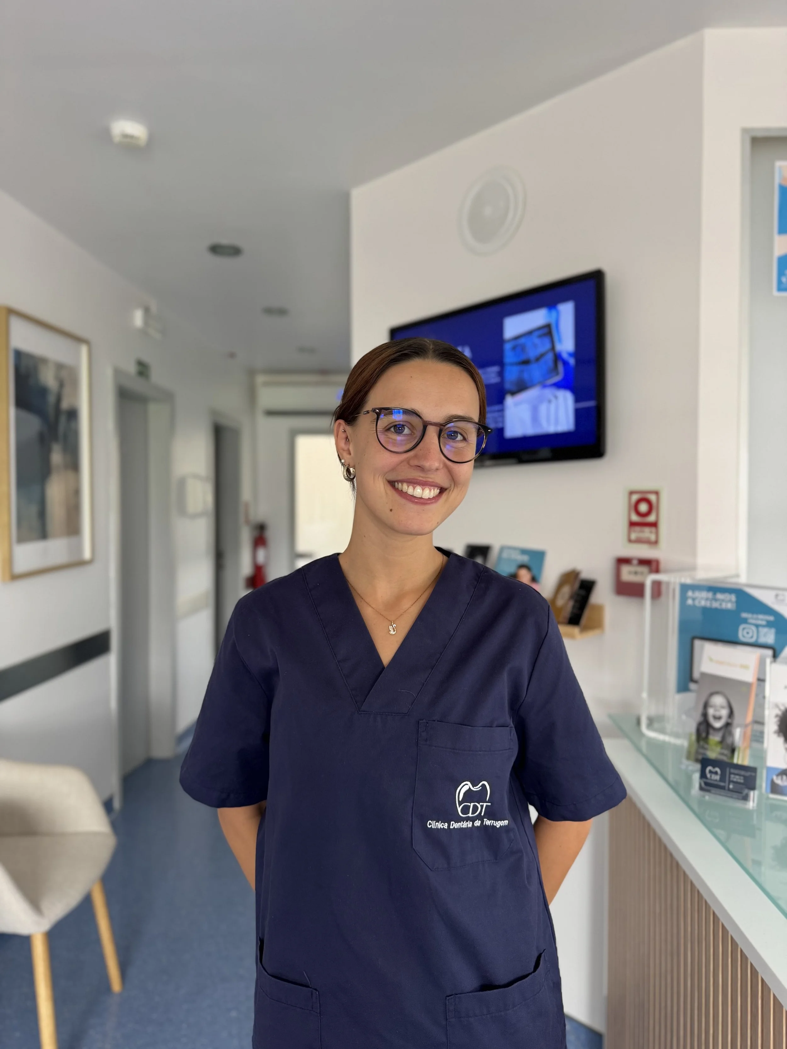 Mulher com cabelo castanho escuro, cabelo liso, usando camisa branca e vestido vinho por baixo, sorrindo, usando brincos pequenos e um colar dourado com pingente