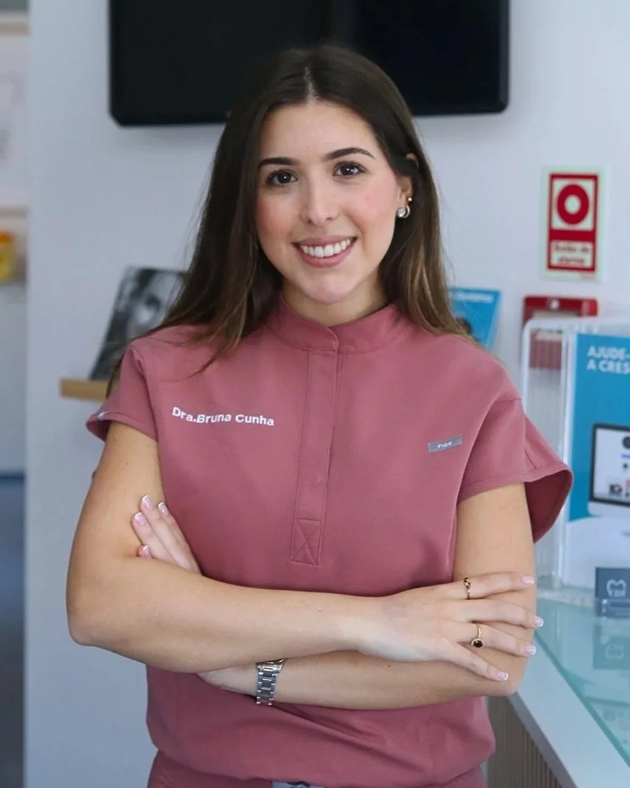 Mulher sorridente com cabelos castanhos claros, usando uniforme rosa com nome "Dra. Bruna Cunha" e acessório de relógio, de braços cruzados, em ambiente de consultório ou loja.