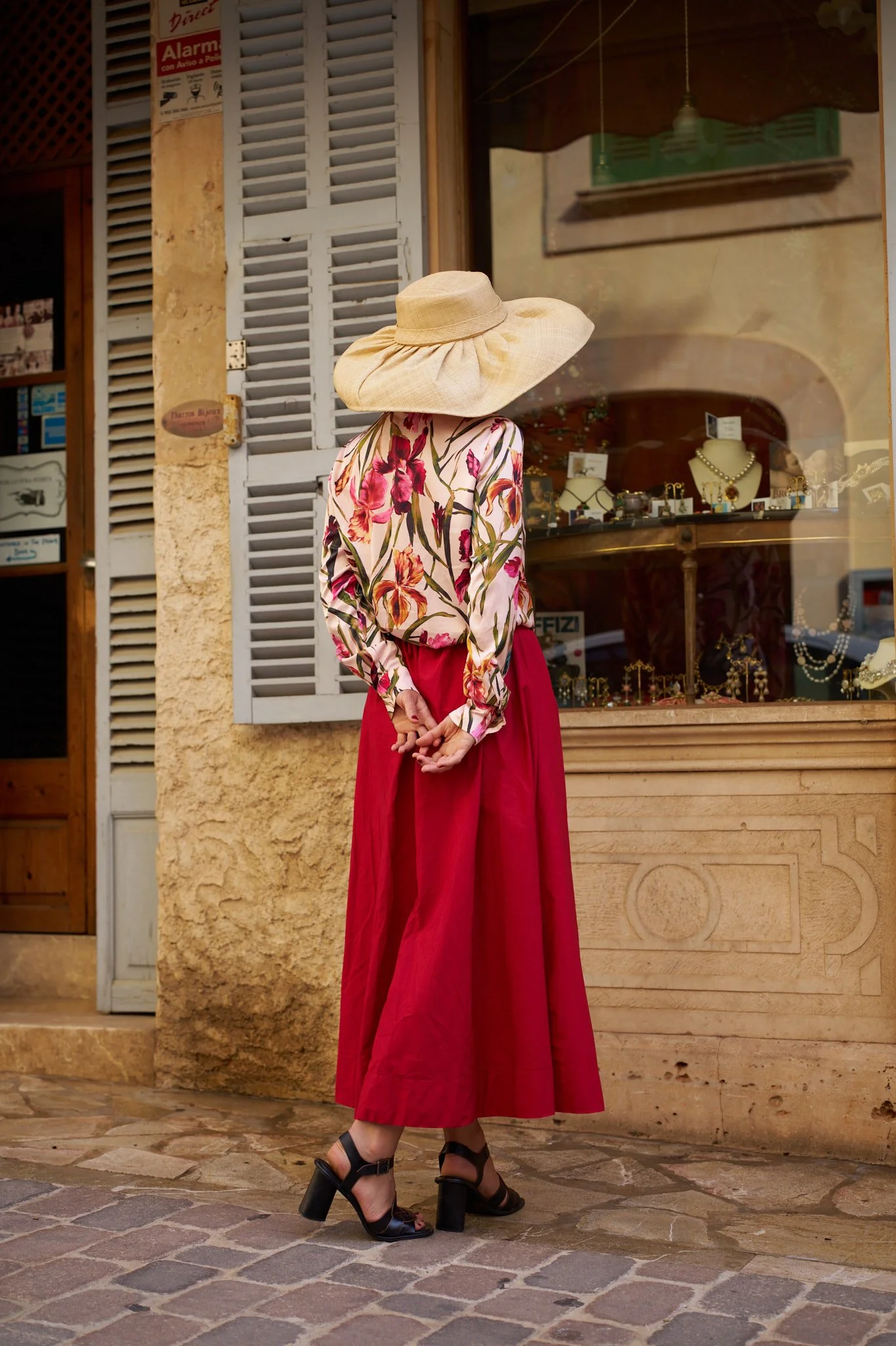 A woman stands on a cobblestone street outside a jewelry store window, wearing a floral blouse, red skirt, black high heels, and a wide-brimmed straw hat.