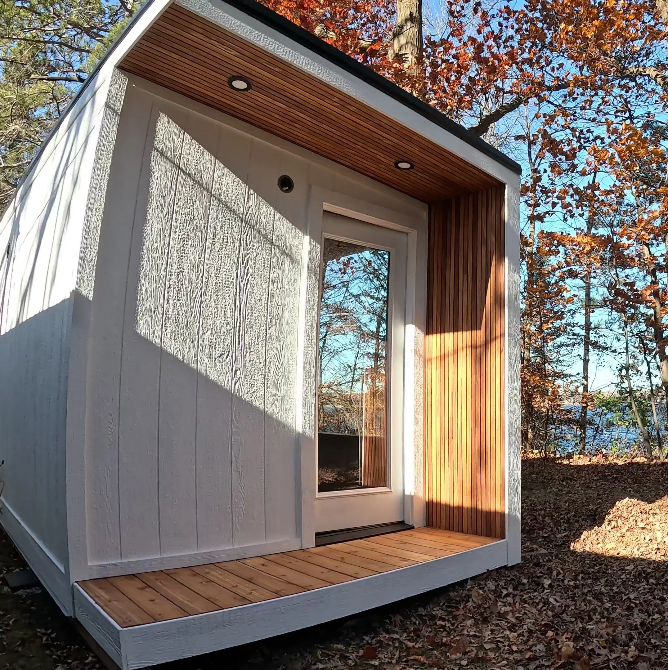 Modern outdoor sauna with a glass door, white exterior walls, and a wooden deck and ceiling, surrounded by autumn trees.