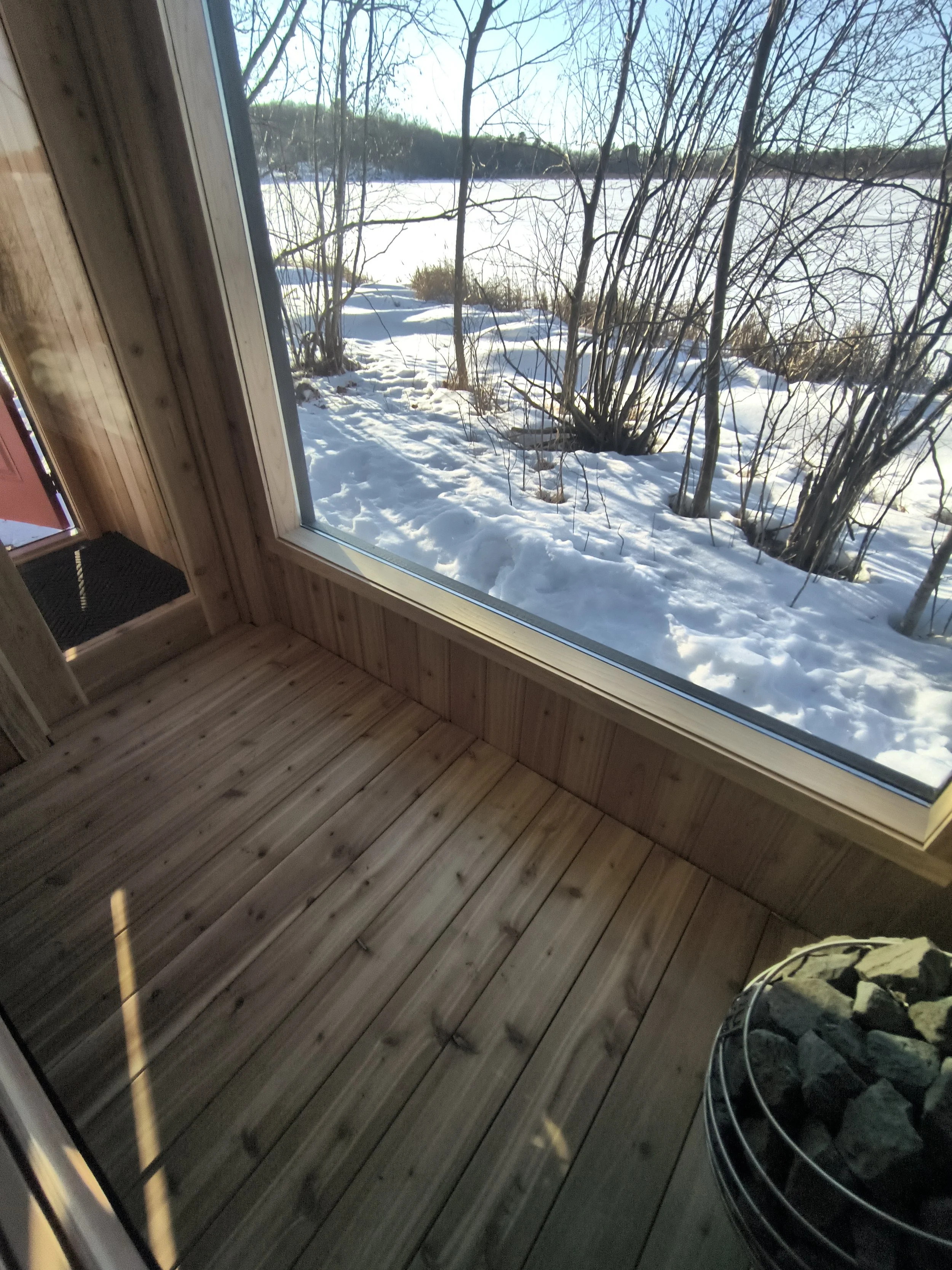 View of snowy landscape through large window, with leafless trees and frozen water outside, inside a wooden sauna with a stone heater.