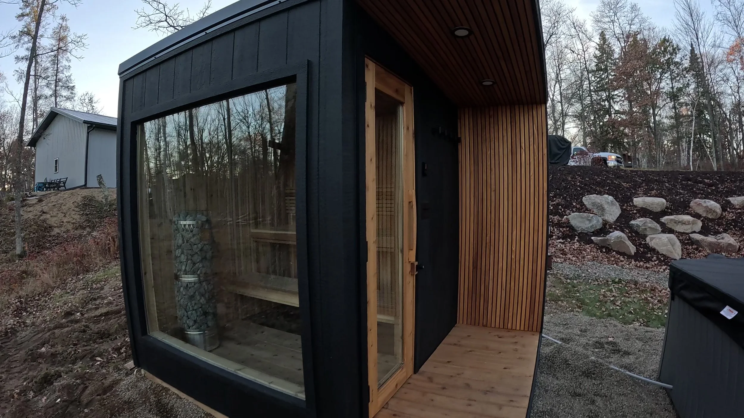 Small modern outdoor sauna with glass door and wooden exterior, located in a wooded area.
