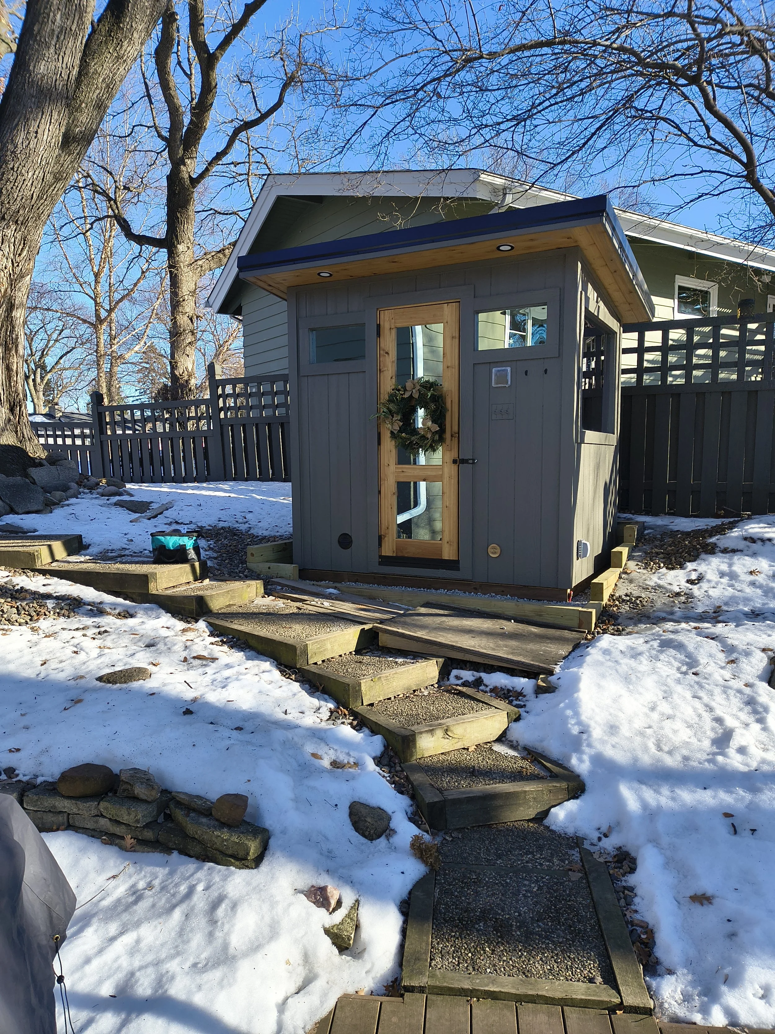 A small gray shed with a wooden door decorated with a Christmas wreath, located in a snowy backyard. There is a stone pathway leading to the shed, bordered by wooden steps and a stone wall, with bare trees and a house with a green exterior in the bac
