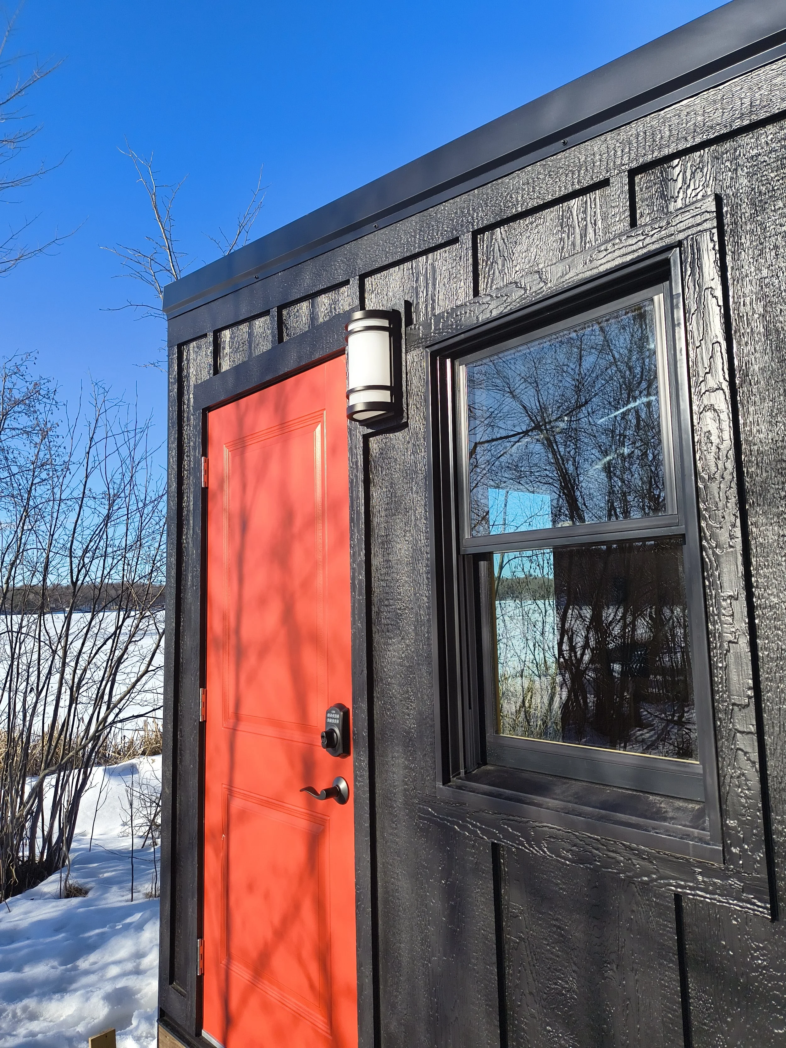 Close-up of a black tiny house with a bright red door, a gray handle, a keypad lock, a black-framed window reflecting trees and blue sky, and a modern light fixture above the door, with snow on the ground and leafless trees in the background.