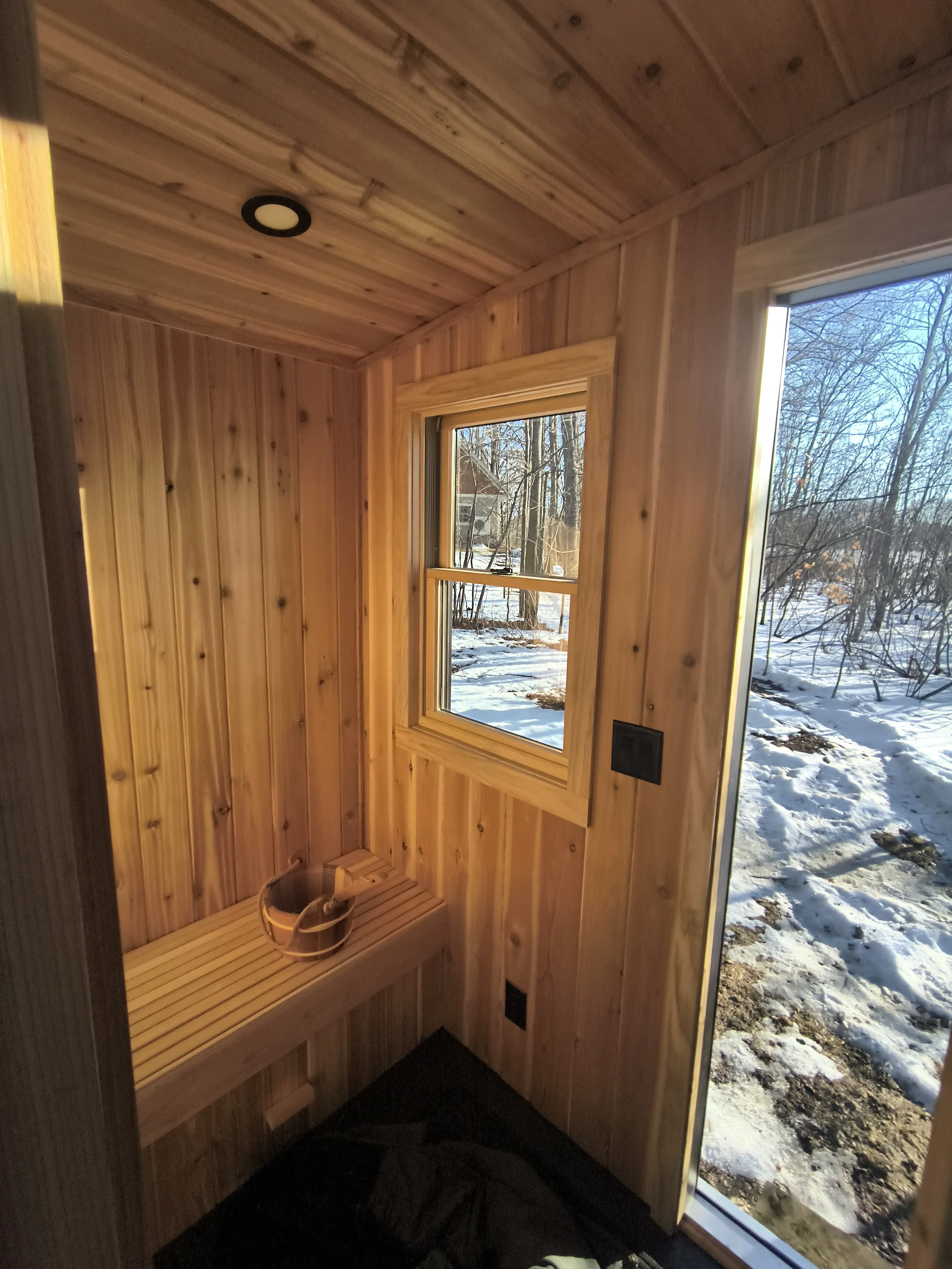 Wood-paneled sauna interior with window, bench, sauna bucket and ladle, and exterior view of snow-covered ground and trees.