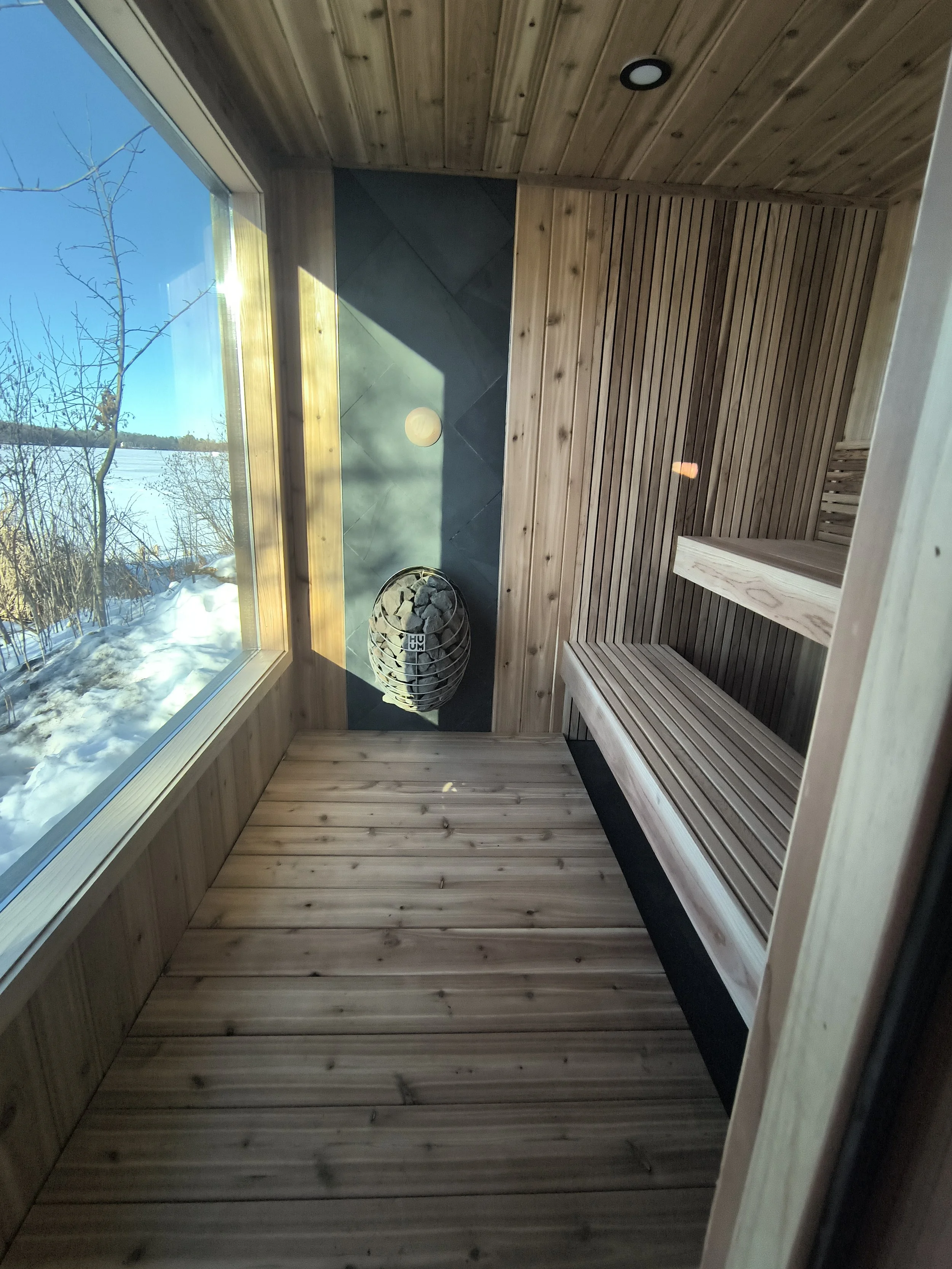 Interior of a wooden sauna with bench seating, a heater with stones, and a large window showing snowy outdoors and trees.