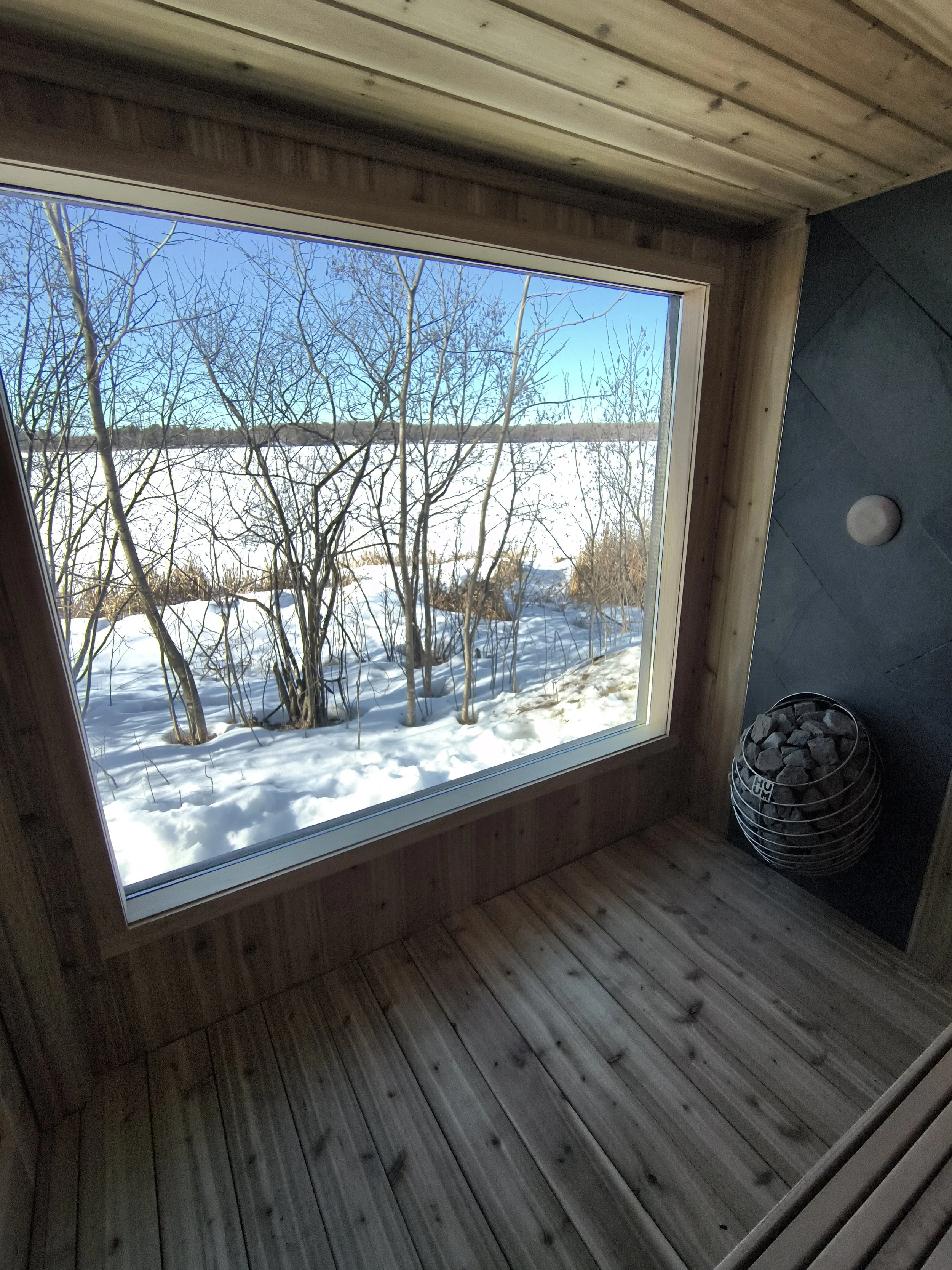 Interior view with a large window showing snow and trees outside, wooden flooring, and a dark wall with a round light and a wire basket filled with rocks.